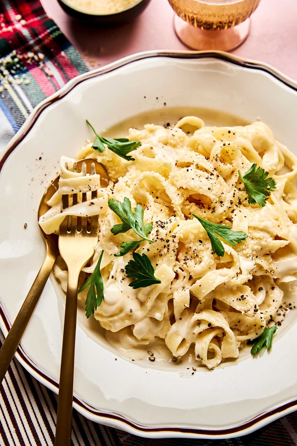 A plate of creamy fettuccine garnished with fresh parsley and black pepper, served with a gold fork and spoon on a white dish with a decorative rim. A patterned napkin and a drink are in the background.