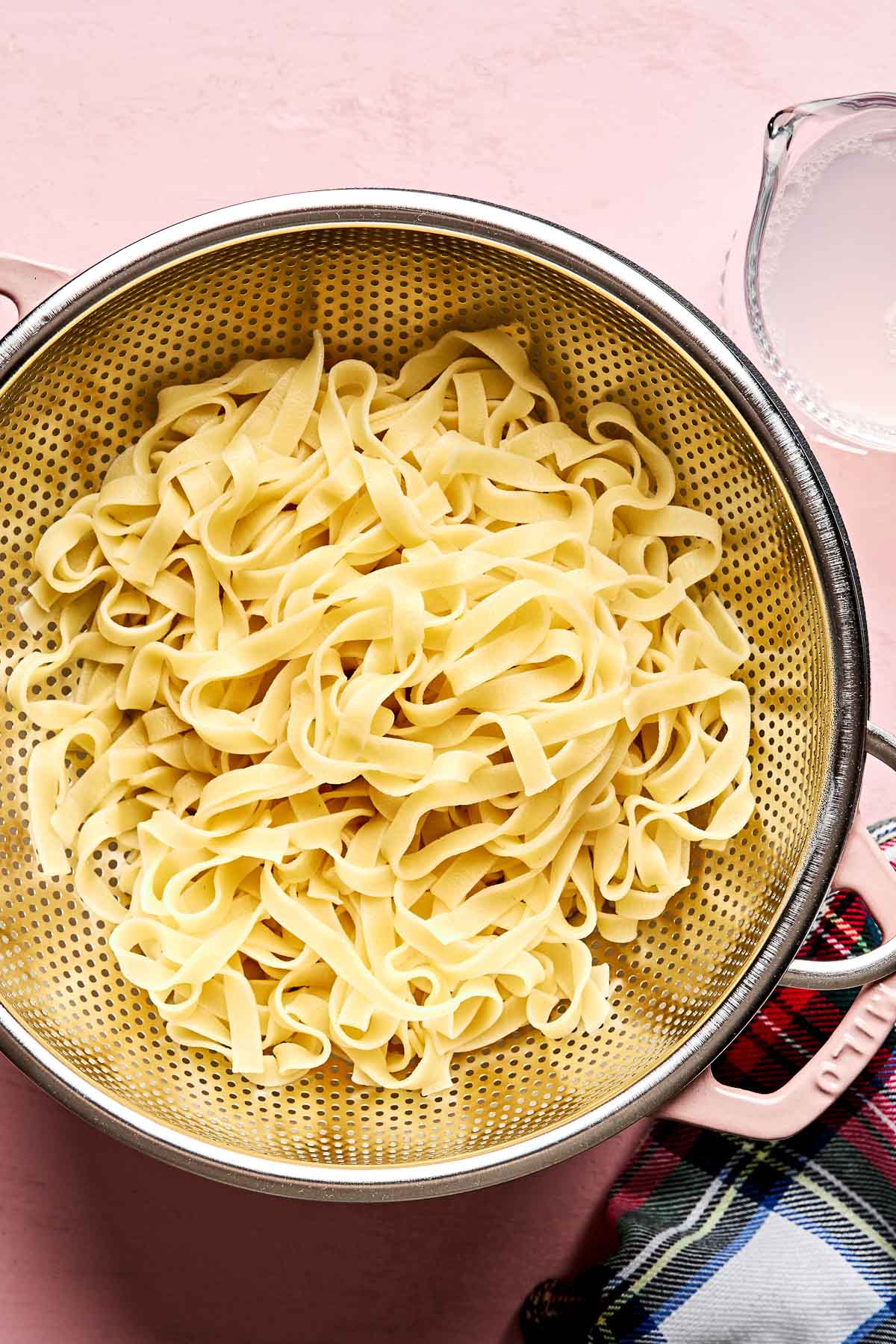 A metal colander filled with cooked fettuccine pasta sits on a pink surface, with a clear measuring cup of pasta water and a plaid cloth nearby.