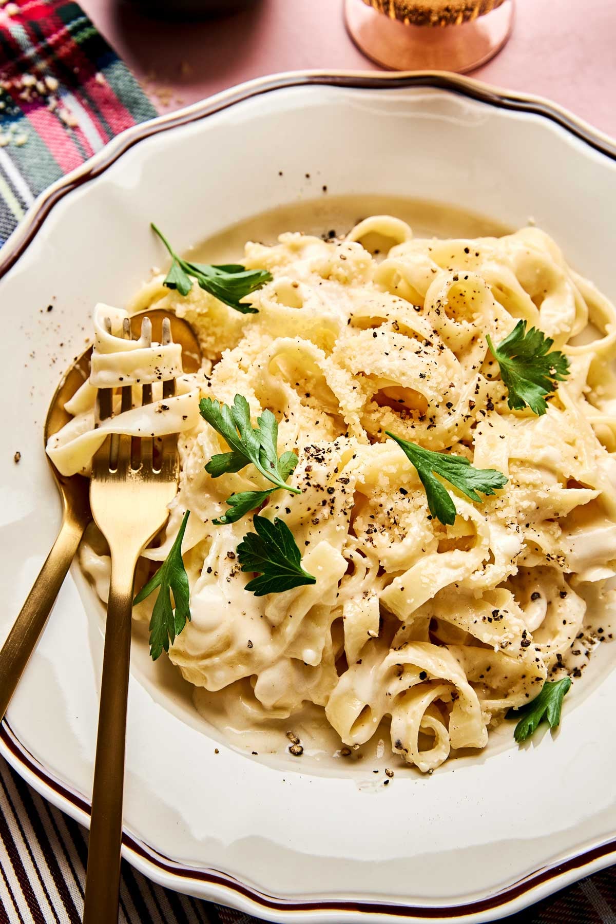 A plate of creamy Christmas fettuccine garnished with fresh parsley and cracked black pepper, served with a gold fork and spoon on a decorative white plate.
