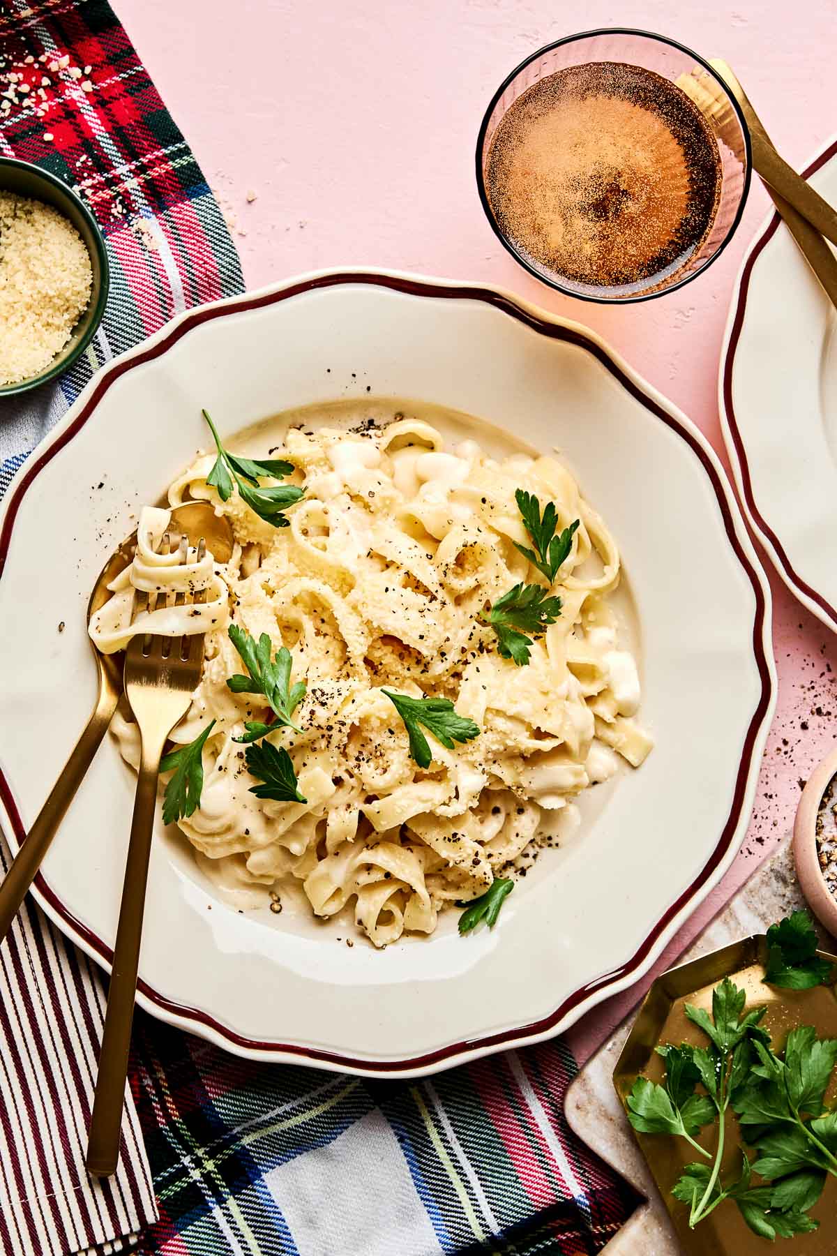 A bowl of creamy Christmas fettuccine garnished with parsley and black pepper sits on a plate with a gold fork. Nearby are a glass of drink, a bowl of grated cheese, and a napkin with a red and blue plaid pattern.