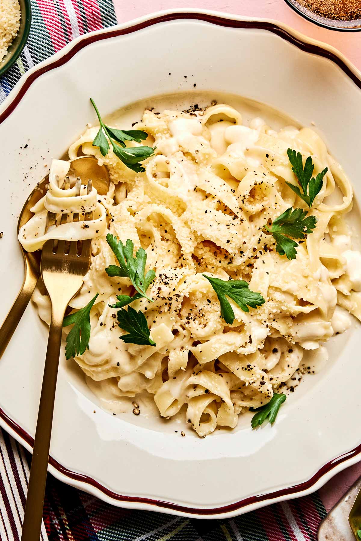 A plate of creamy Christmas fettuccine garnished with fresh parsley and sprinkled with black pepper, served with a gold fork and spoon on a patterned cloth.