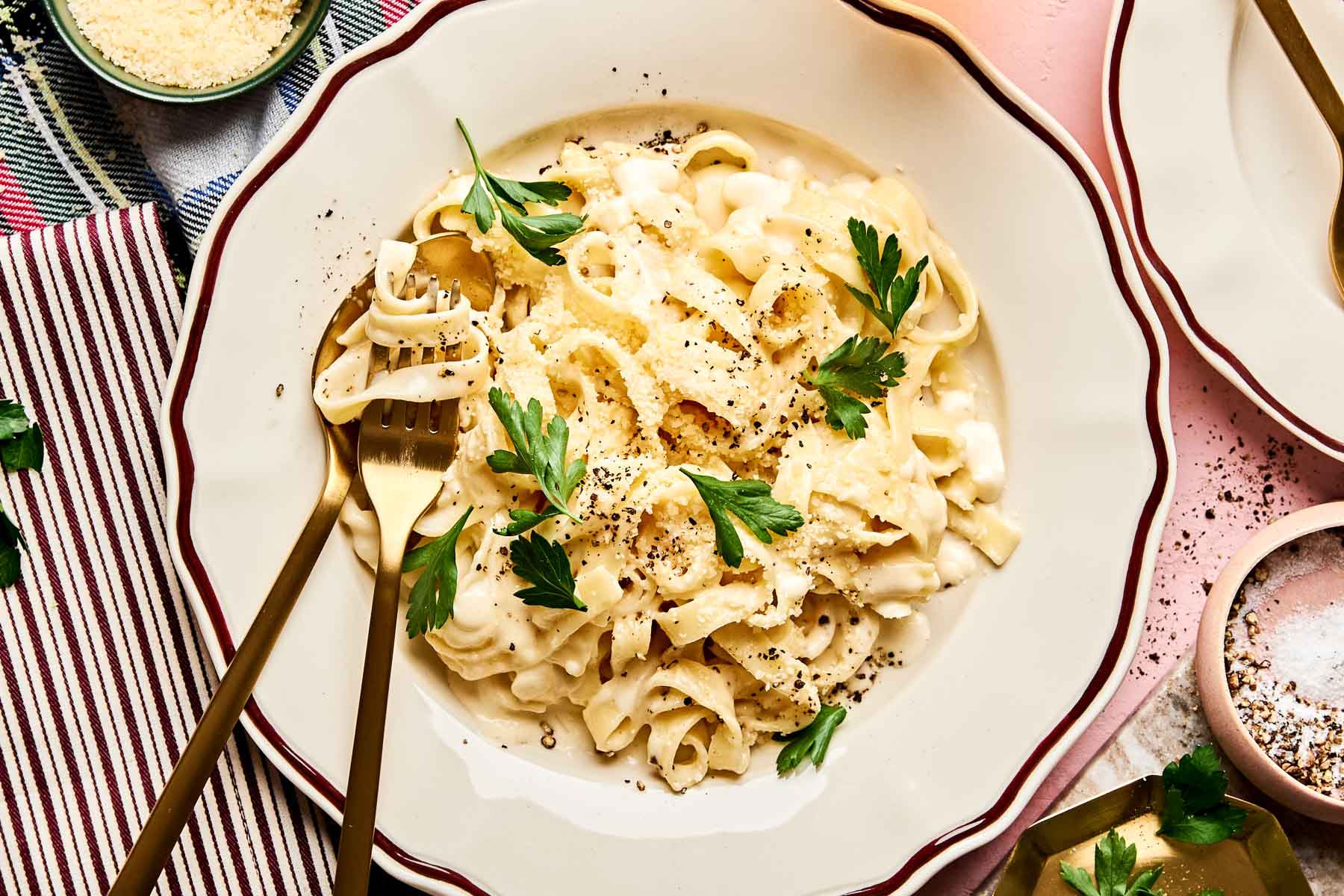 A plate of creamy Christmas fettuccine garnished with parsley and cracked black pepper, served with two gold-colored forks, and surrounded by small bowls of grated cheese and spices.