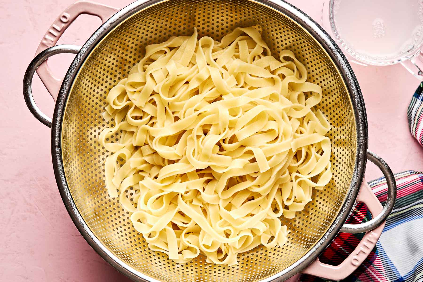 A metal colander filled with cooked fettuccine pasta sits on a pink surface next to a colorful towel and a glass bowl of pasta water.