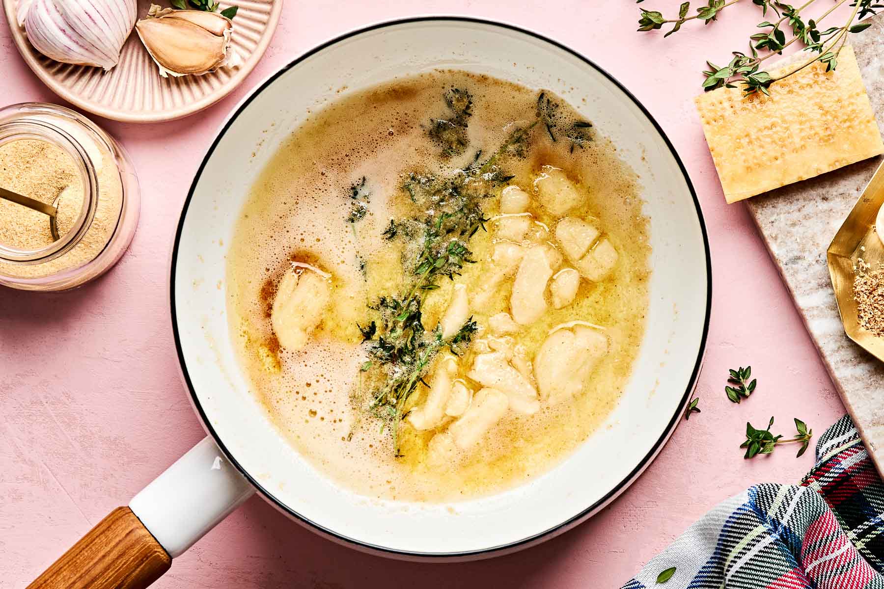A white pan with melted butter, garlic cloves, and fresh thyme sprigs sits on a pink surface near grated cheese, a garlic bulb, a jar of seasoning, and a colorful cloth.