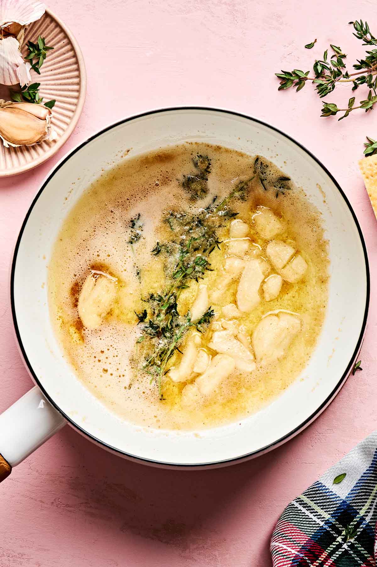 A white pan contains melted butter with whole garlic cloves and sprigs of fresh thyme. There is a plate with garlic cloves and a cloth napkin nearby, all on a pink surface.