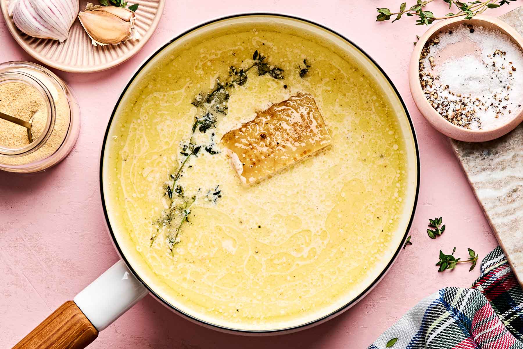 A white saucepan filled with creamy sauce, topped with a chunk of parmesan cheese and fresh thyme sprigs, sits on a pink surface. Surrounding it are garlic cloves, a salt bowl, a spice jar, and a plaid cloth.