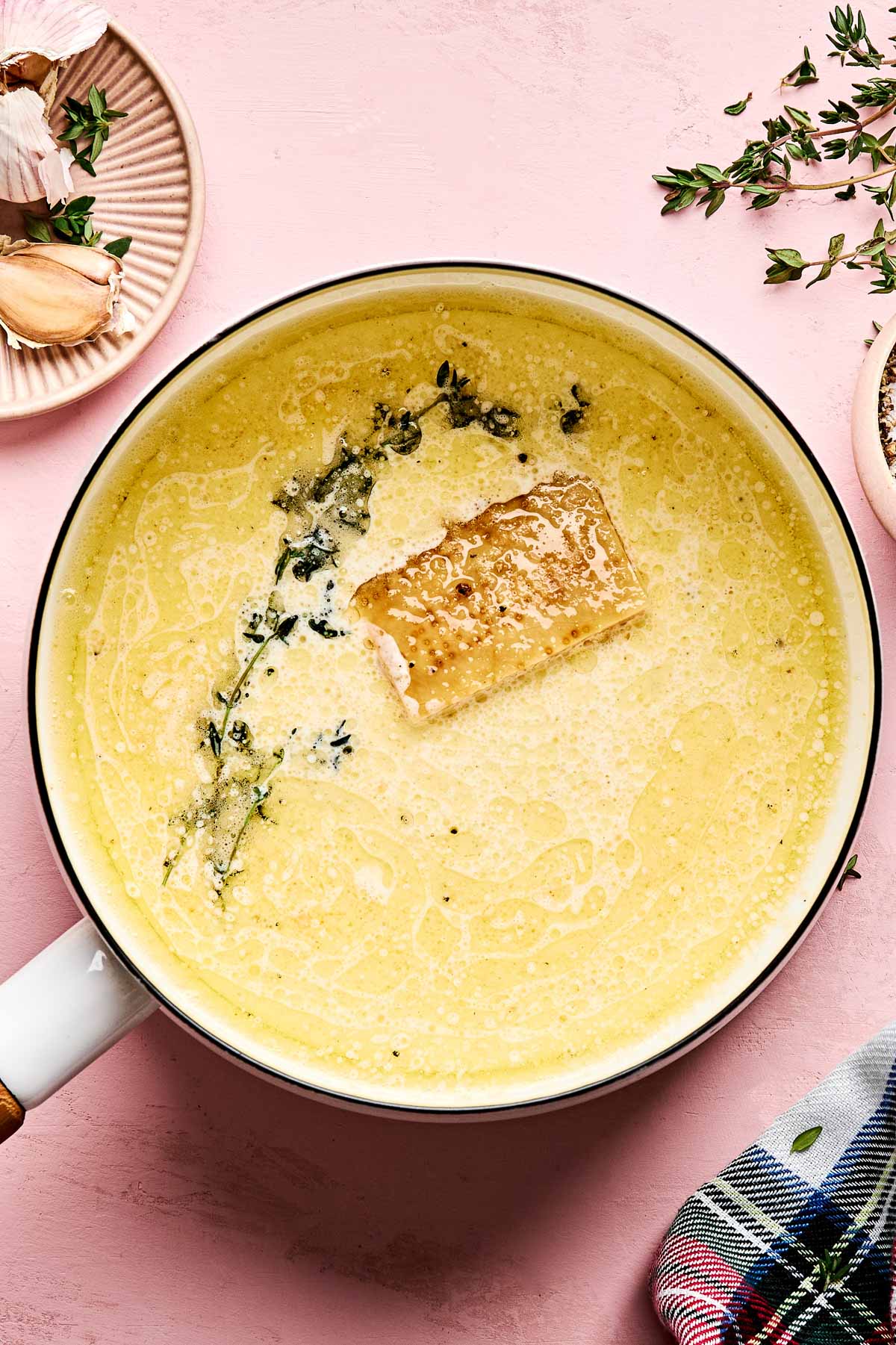 A creamy pasta sauce in a white saucepan, garnished with herbs and a chunk of Parmesan cheese. Fresh thyme and garlic are nearby on the pink tabletop, with a checkered cloth partly visible in the corner.