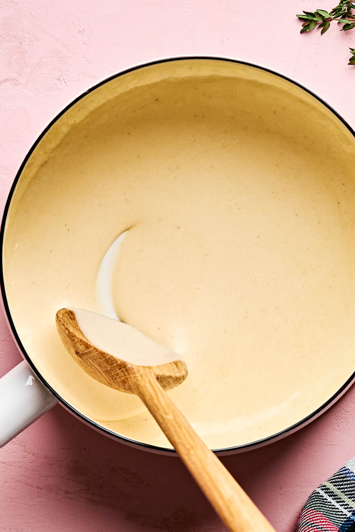 A pot filled with creamy pasta sauce sits on a pink surface, with a wooden spoon resting inside and some herbs visible in the upper right corner.