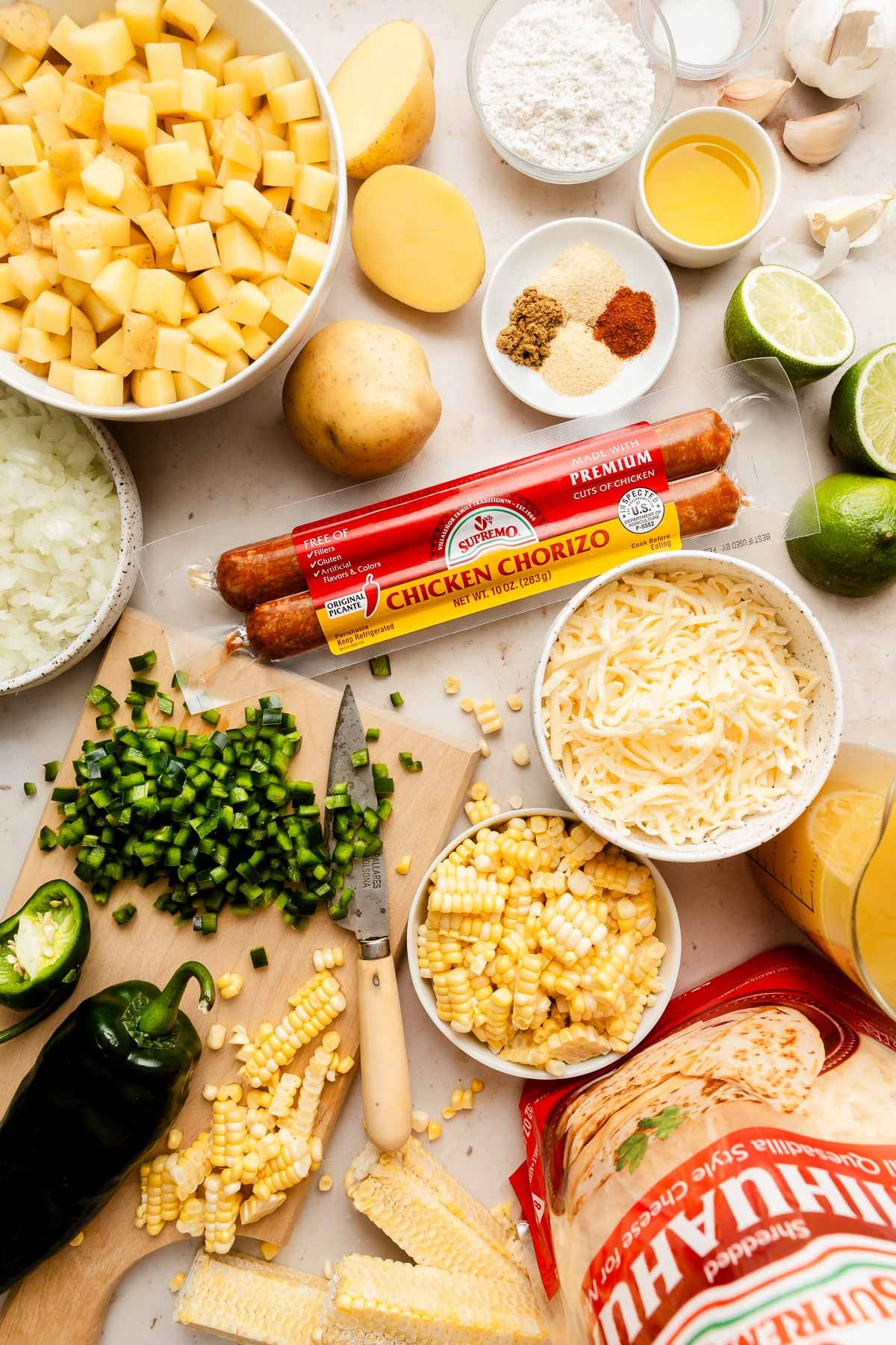 An overhead view of diced potatoes, chopped onions, jalapeños, corn, shredded cheese, flour, spices, limes, garlic, and chicken chorizo sausage arranged on a kitchen counter with tortillas.