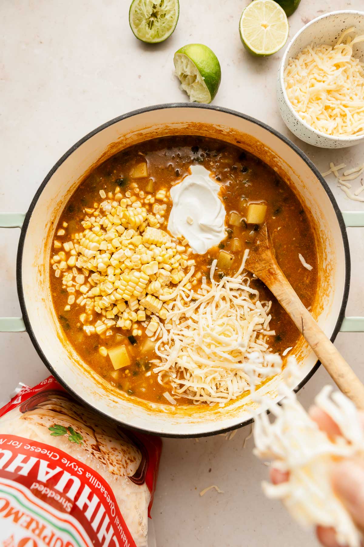 Overhead view of a pot of corn chowder with corn kernels, shredded cheese, and a dollop of sour cream on top, next to a bowl and a bag of cheese, with halved limes nearby. A wooden spoon rests in the pot.