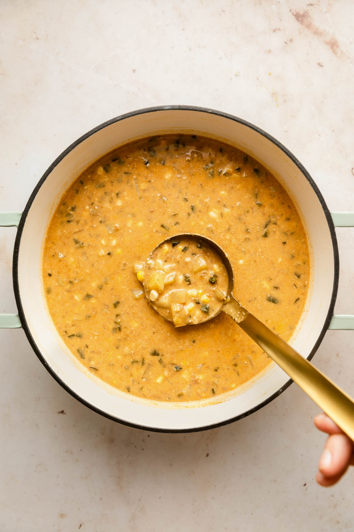 A hand holds a ladle scooping creamy, poblano corn chowder with visible chunks of vegetables and herbs from a white pot on a light-colored countertop.