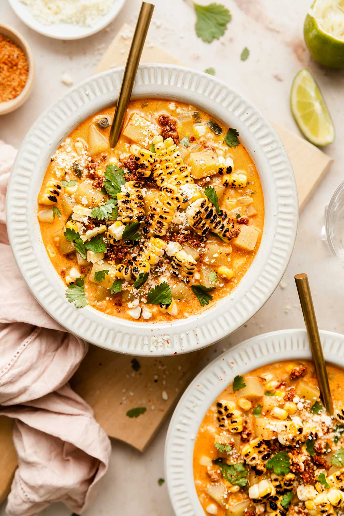 Two white bowls filled with creamy poblano corn chower topped with grilled corn, diced vegetables, crumbled cheese, chopped cilantro, and chili powder, each with a gold spoon. A lime wedge and cloth napkin are nearby on the table.