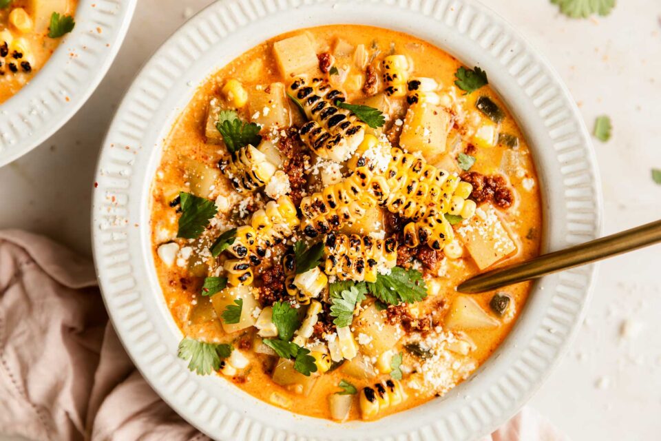 A bowl of creamy poblano corn chowder with chorizo topped with grilled corn, chopped cilantro, crumbled cheese, and a sprinkle of spices. A gold spoon rests in the bowl, and a light napkin is nearby on the table.