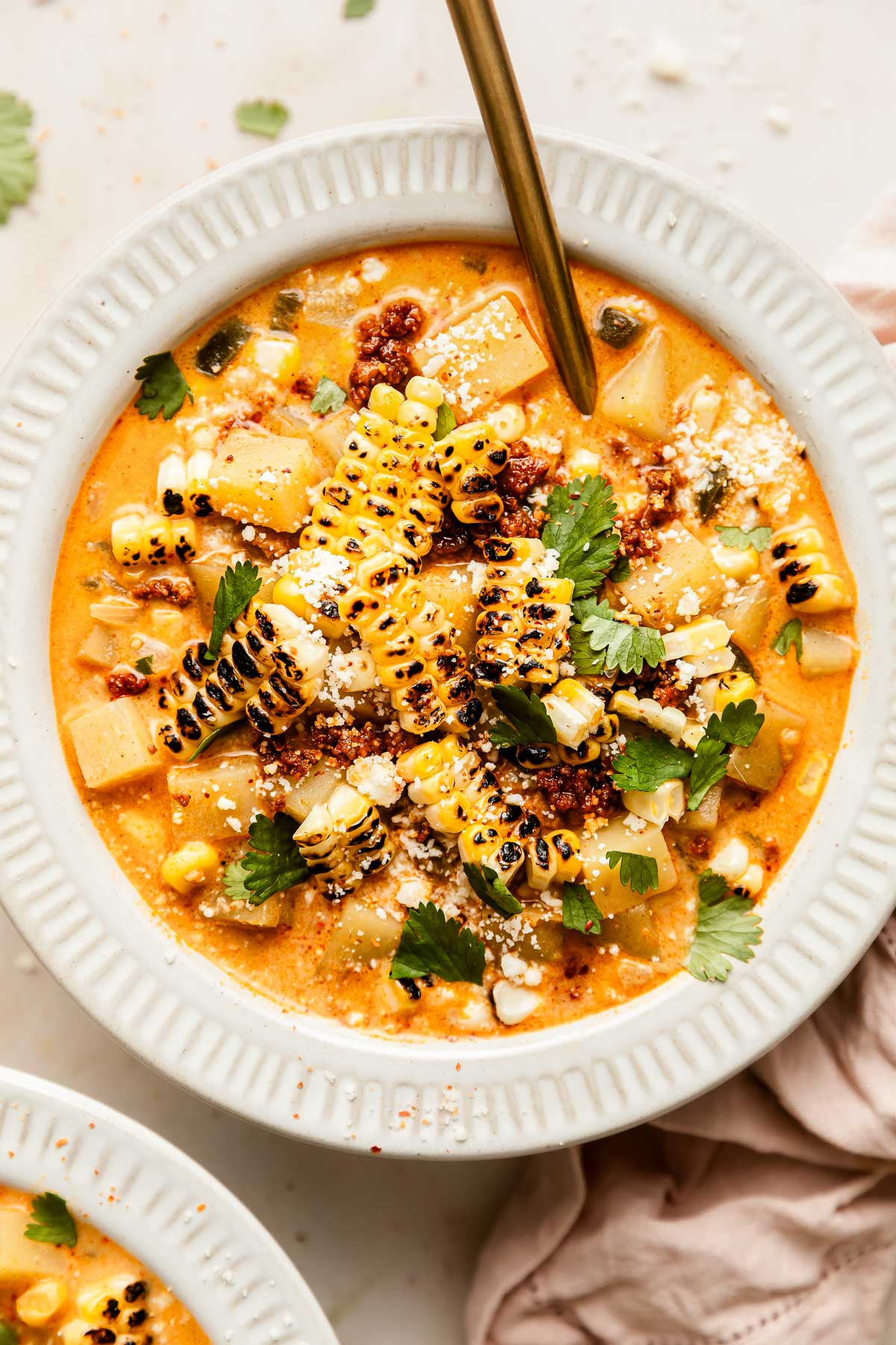 A bowl of creamy poblano corn chowder with chorizo topped with grilled corn, chopped cilantro, crumbled cheese, and spices, with a spoon inside. Cubed vegetables are visible in the soup, and a pink napkin lies nearby.