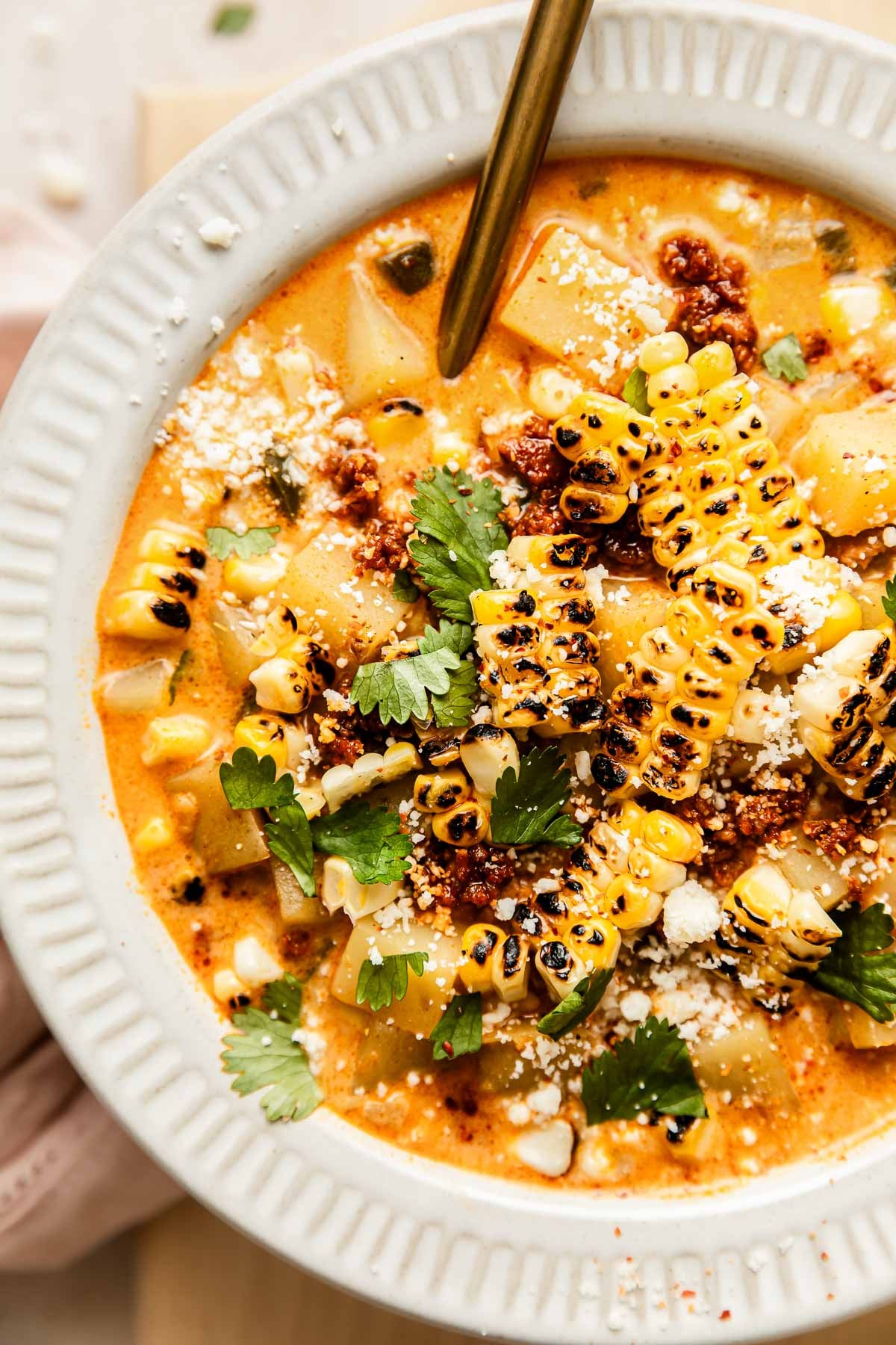 A close-up of a bowl of creamy corn chowder with poblano peppers and chorizo topped with grilled corn, potatoes, fresh cilantro, crumbled cheese, and a sprinkle of spices, with a spoon resting inside the bowl.