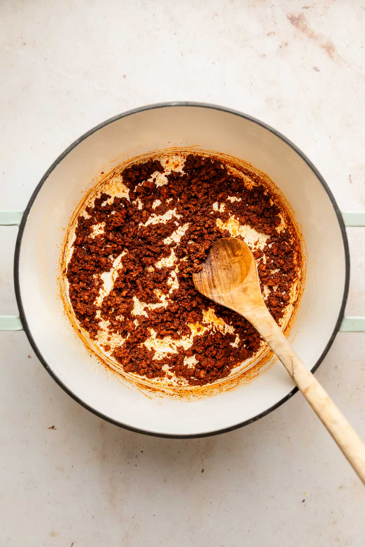 A white pot with cooked, browned ground chorizo sits on a light surface. A wooden spoon rests inside the pot, partially covered in the meat mixture.