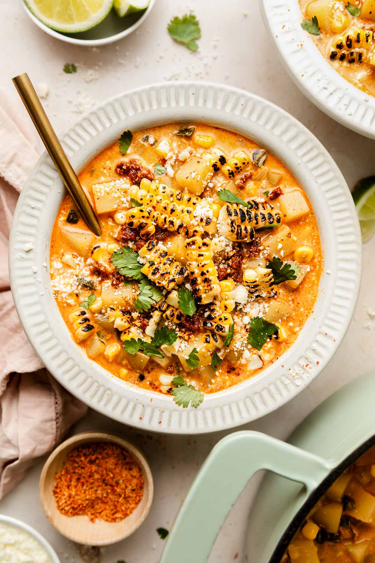 A bowl of creamy corn chowder topped with grilled corn, crumbled cheese, fresh cilantro, and chili flakes, with a gold spoon. Lime wedges, seasoning, and another bowl of soup are nearby on a light surface.