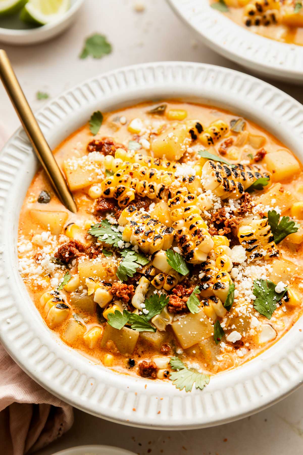 A bowl of creamy corn chowder topped with grilled corn, crumbled cheese, fresh cilantro, and pieces of sausage, served with a spoon on the side. Lime wedges and another bowl are visible in the background.