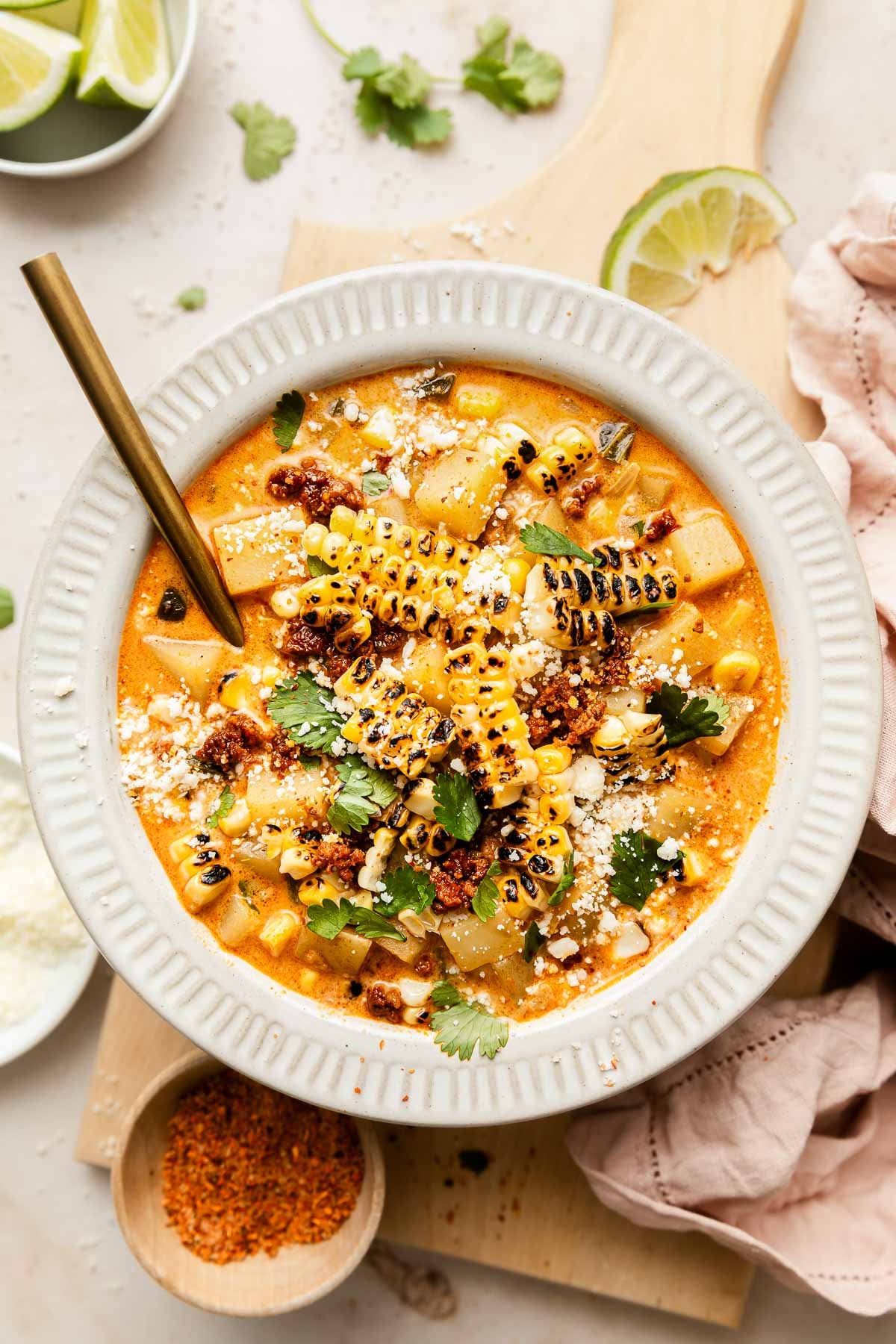 A bowl of creamy poblano corn chowder topped with grilled corn, crumbled cheese, fresh cilantro, and a sprinkle of spices. A gold spoon rests in the bowl, with lime wedges and a small dish of seasoning nearby.