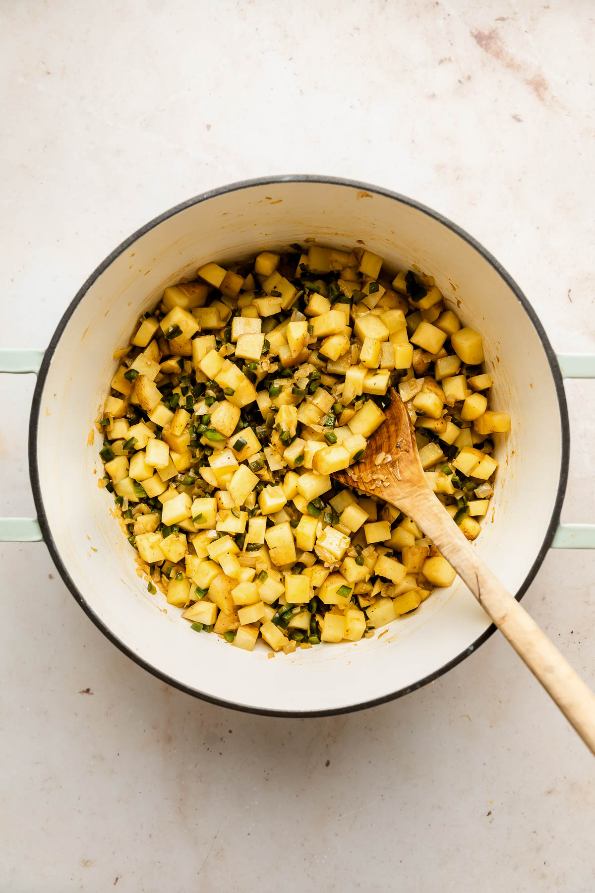 Diced potaties being cooked in a white pot with green handles, mixed with herbs, and stirred with a wooden spoon on a light countertop.