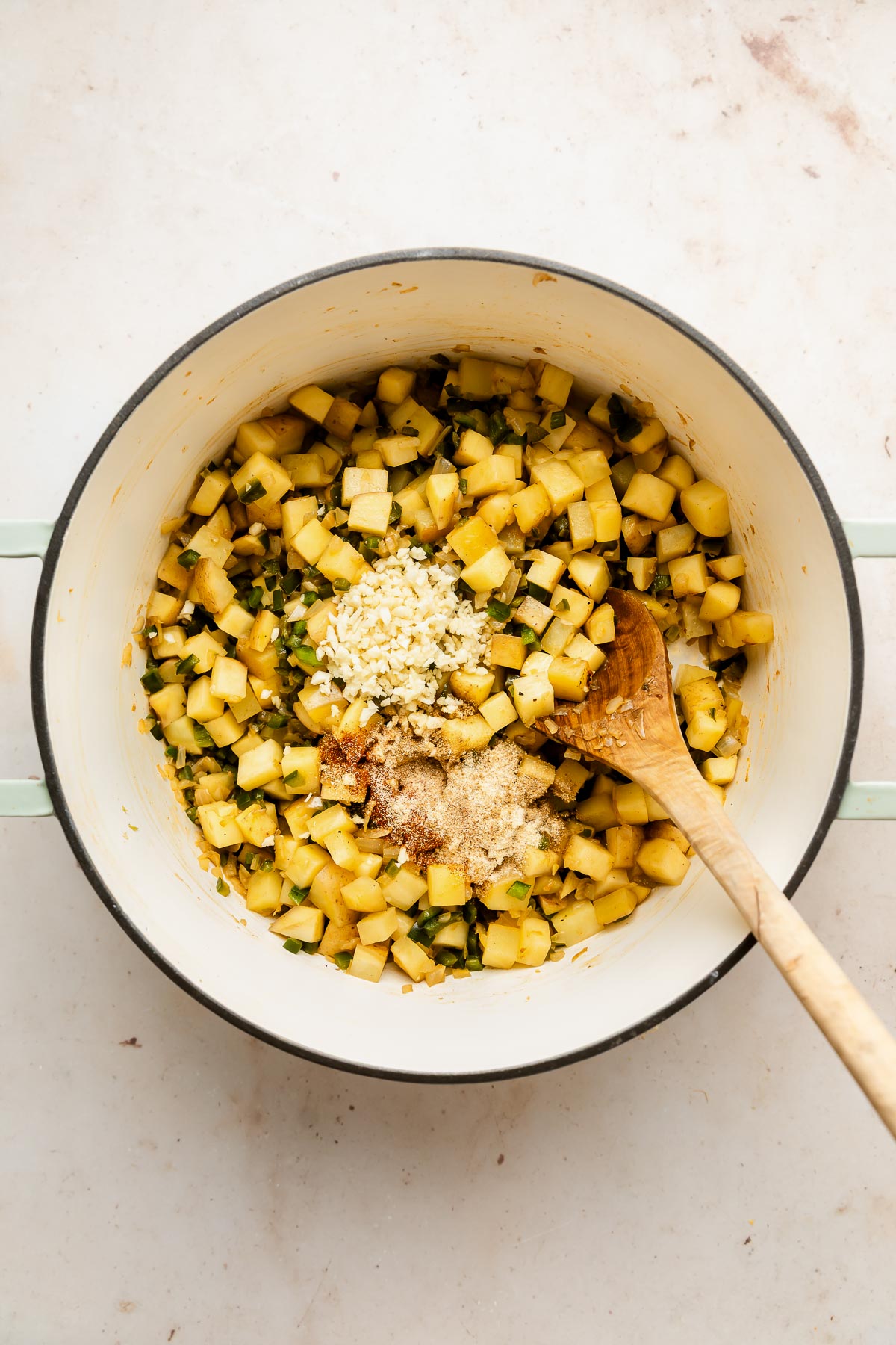 A large white pot contains diced yellow potatoes, chopped garlic, and brown sugar, being mixed with a wooden spoon on a light-colored surface.
