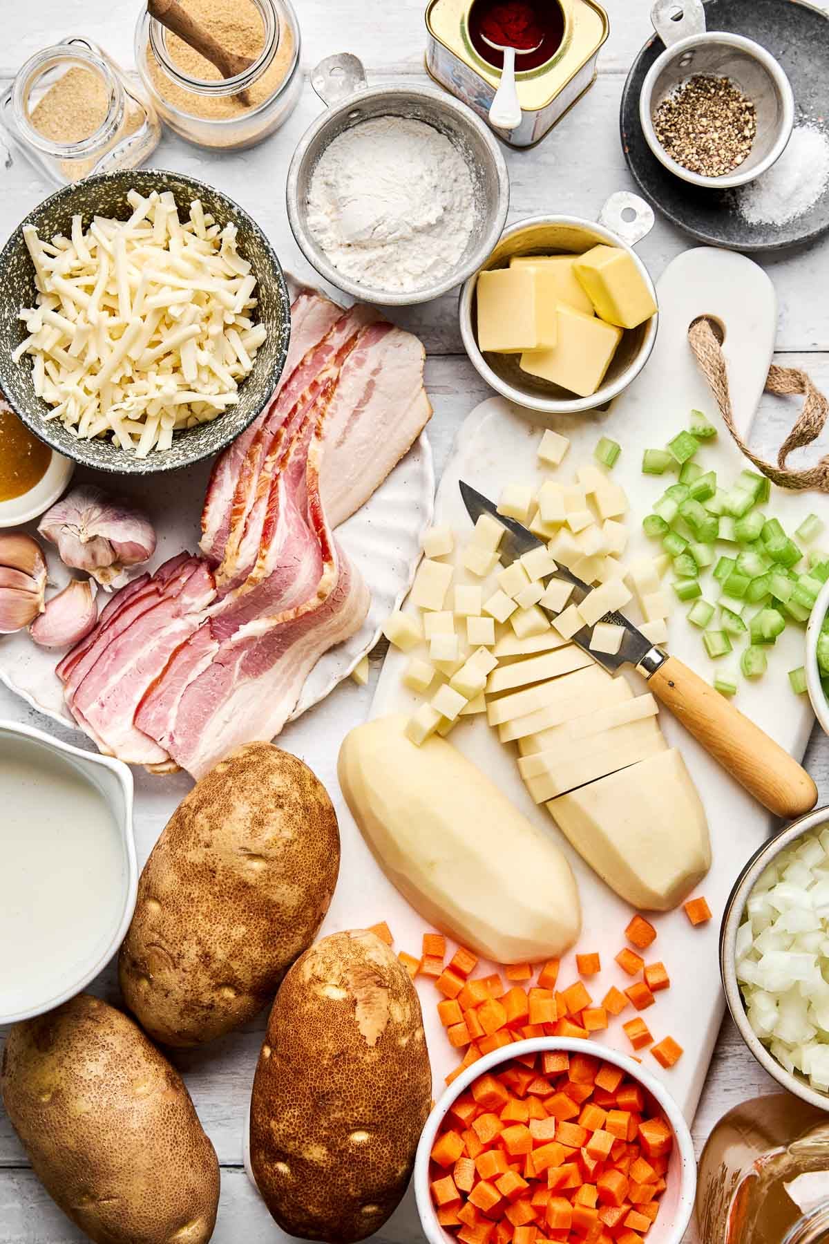 Top-down view of ingredients on a white surface, including peeled and sliced potatoes, bacon, grated cheese, chopped carrots, celery, onions, butter, flour, milk, garlic, and spices.