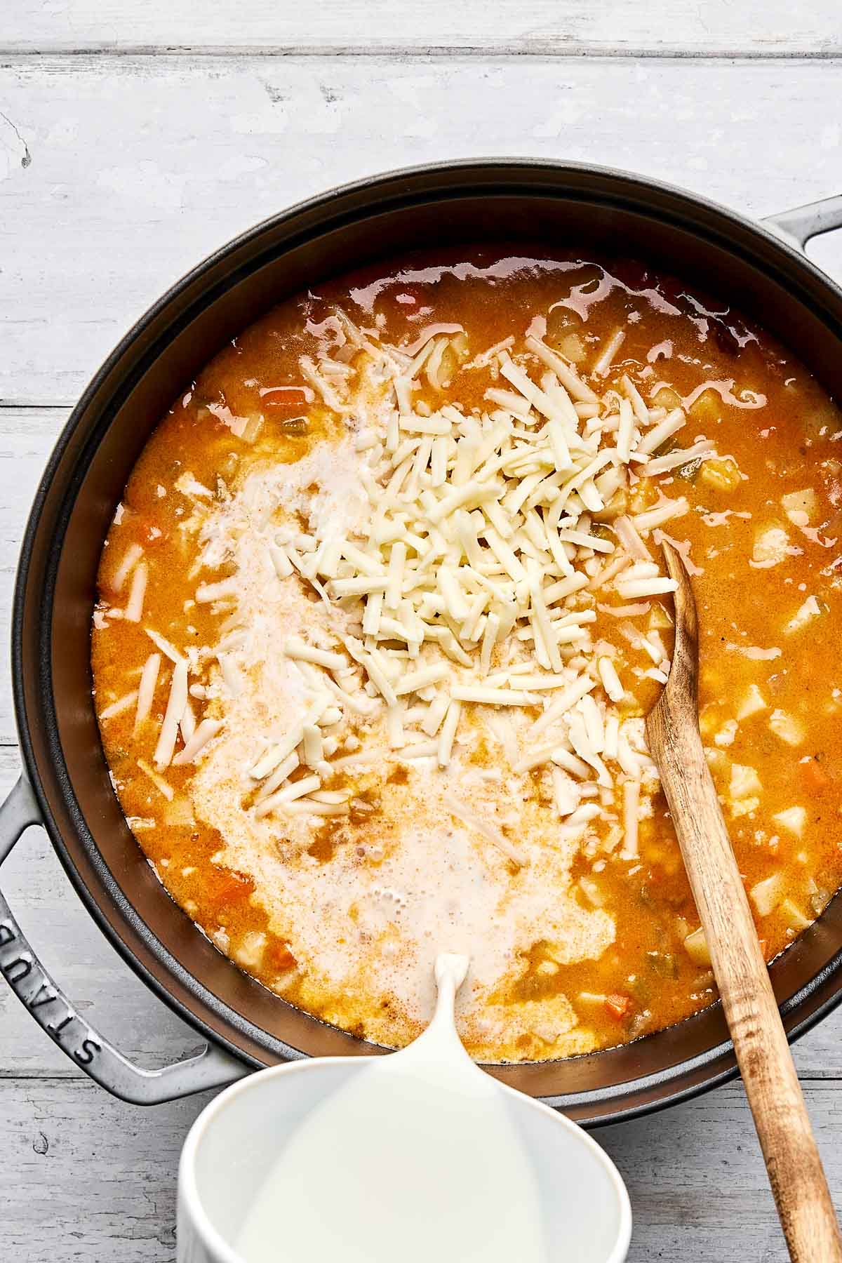 A pot of potato soup on a white wooden surface, topped with shredded cheese and being poured with milk from a white jug, with a wooden spoon resting inside the pot.