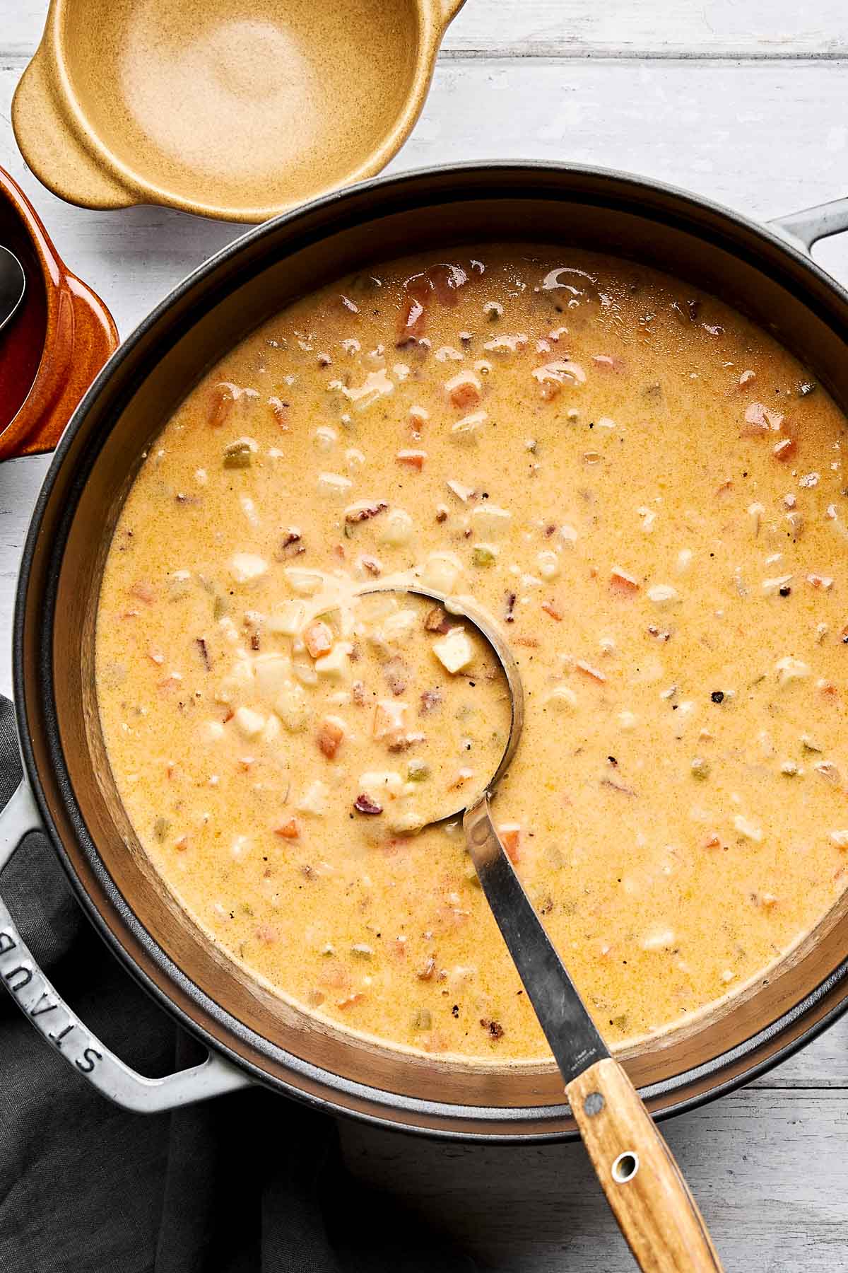 A pot of creamy potato soup with visible chunks of vegetables and bacon, with a metal ladle resting inside. The soup is thick and hearty. An empty bowl and cloth are nearby on a light surface.