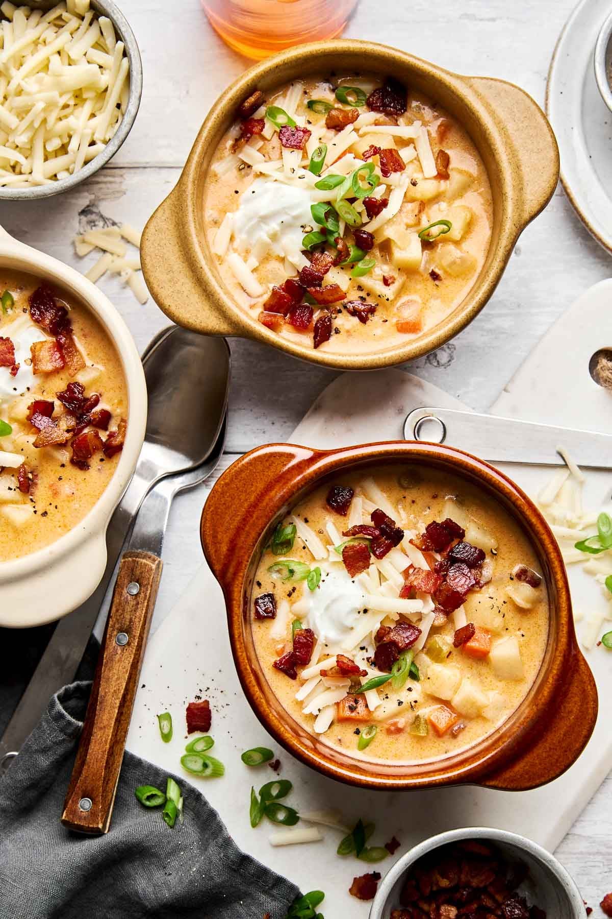 Two bowls of creamy potato soup topped with shredded cheese, bacon bits, sour cream, and chopped green onions, surrounded by a spoon, a napkin, and small dishes of extra toppings on a white surface.