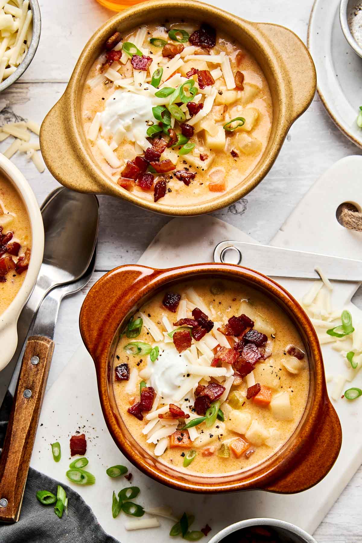 Two bowls of creamy potato soup topped with shredded cheese, crispy bacon, a dollop of sour cream, and chopped green onions, placed on a light surface with spoons and extra garnishes nearby.