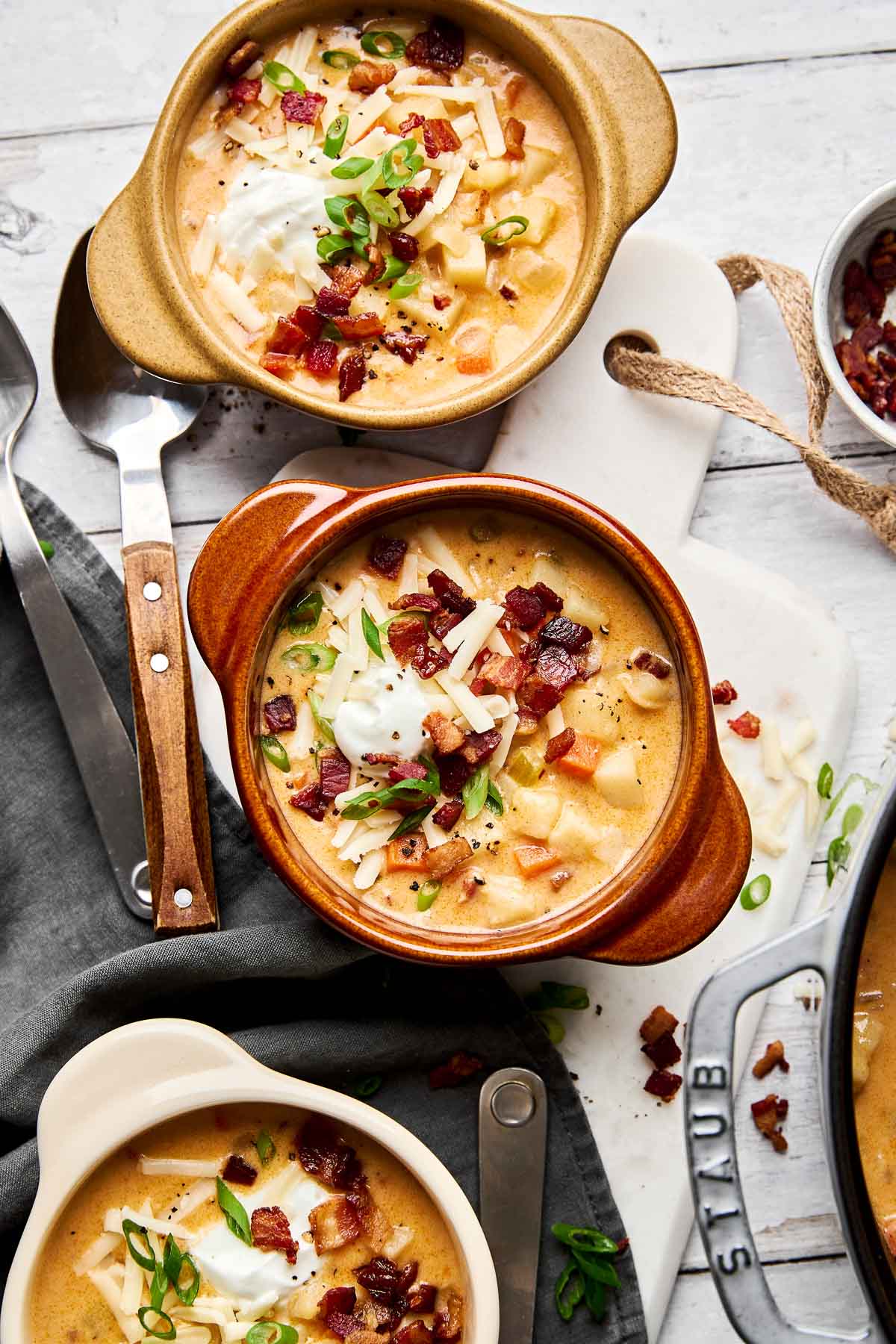 Three bowls of creamy potato soup topped with shredded cheese, sour cream, crispy bacon pieces, and chopped green onions, arranged on a white wooden surface with spoons and a gray napkin nearby.