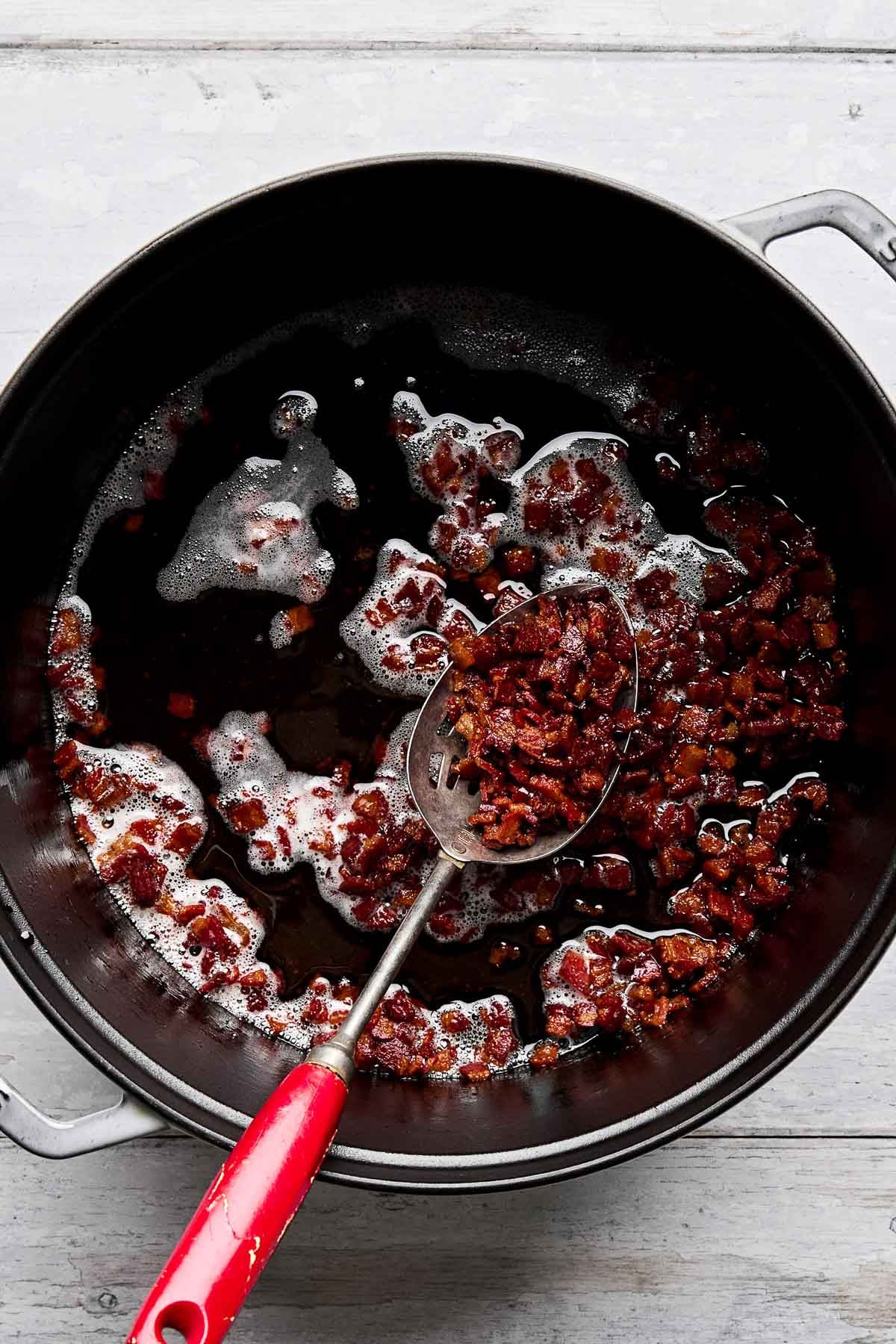 A black pot with cooked, crispy bacon bits and some sizzling grease. A slotted spoon with a red handle is resting in the pot, holding a portion of the bacon bits. The pot is on a light gray wooden surface.