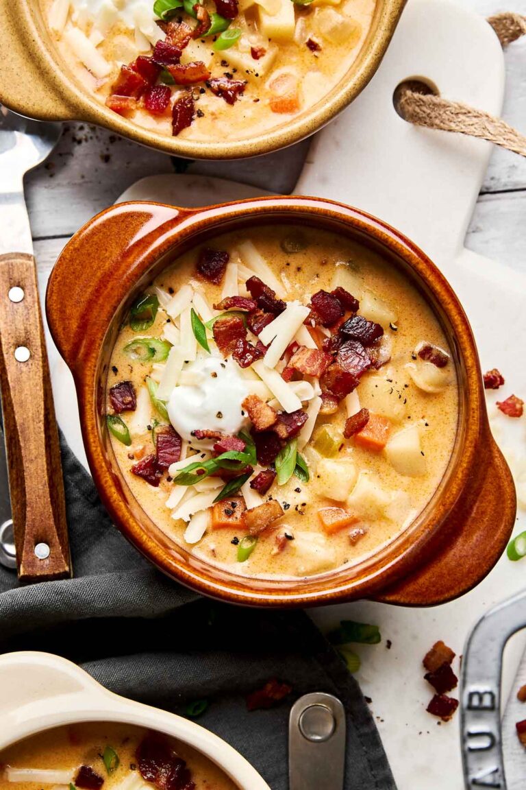 A bowl of creamy potato soup topped with shredded cheese, crispy bacon bits, chopped green onions, and a dollop of sour cream, served in a brown ceramic bowl on a white surface with a knife nearby.