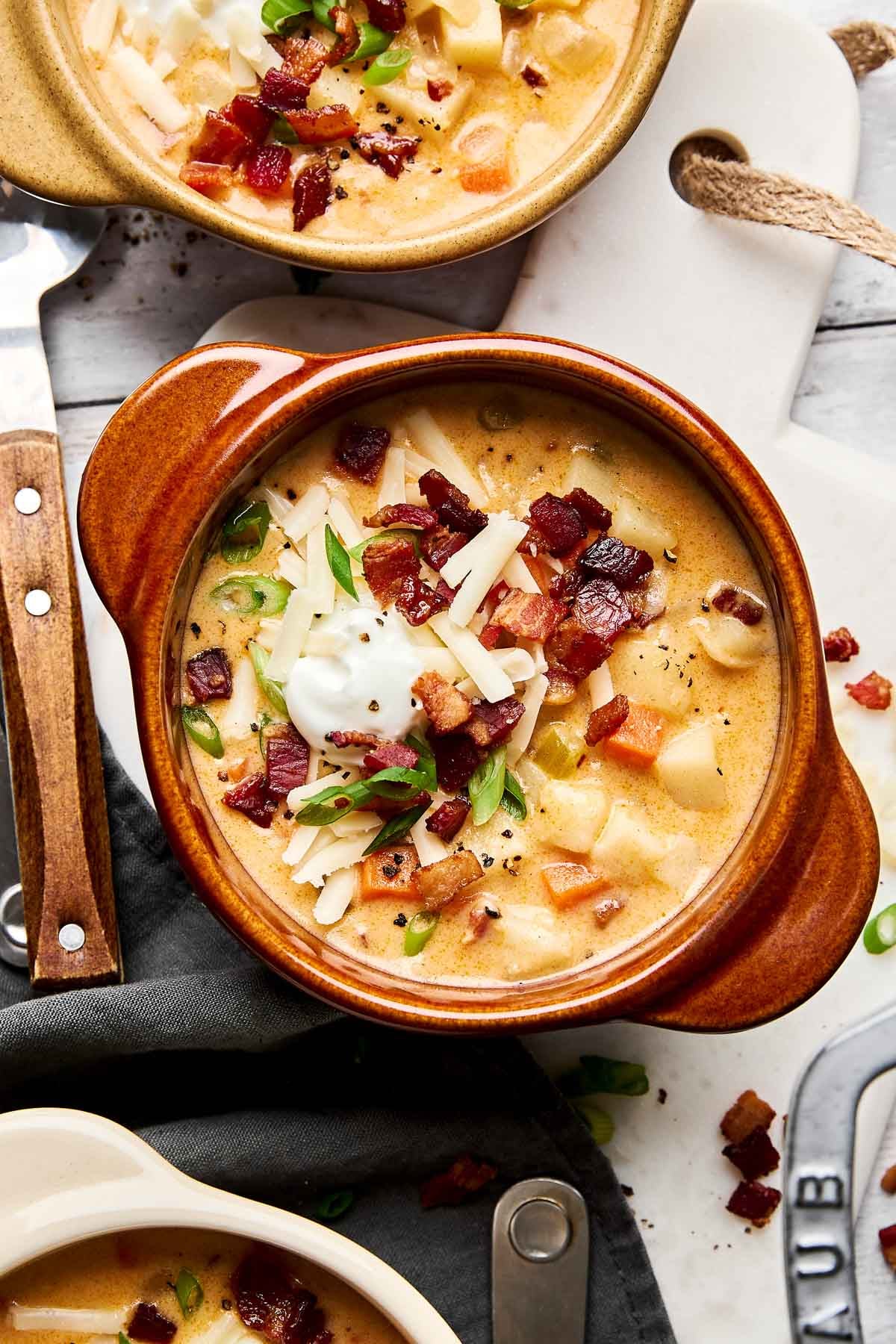A bowl of creamy potato soup topped with shredded cheese, crispy bacon bits, chopped green onions, and a dollop of sour cream, served in a brown ceramic bowl on a white surface with a knife nearby.
