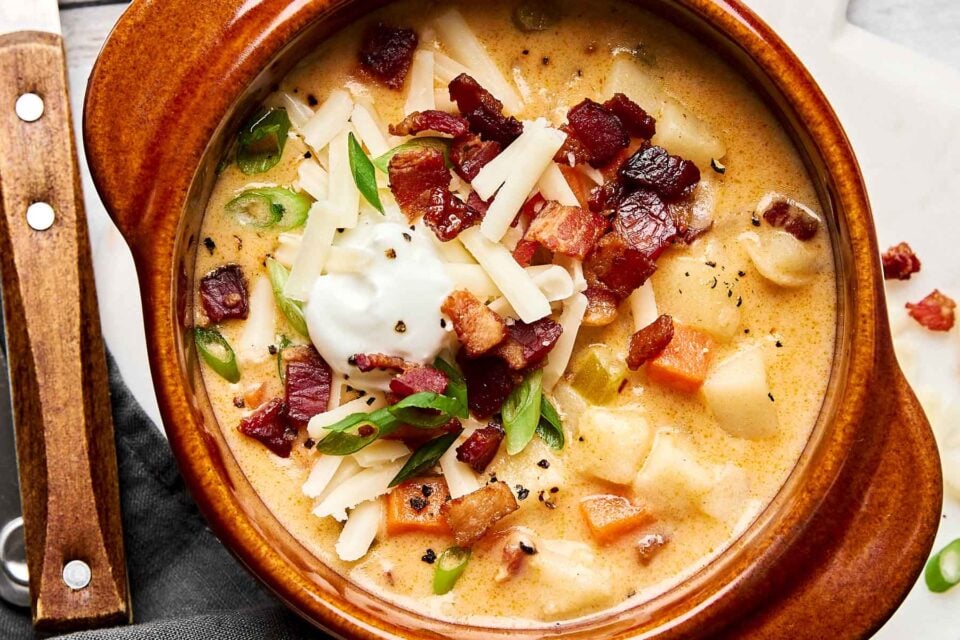 A bowl of creamy potato soup topped with crispy bacon, shredded cheese, sliced green onions, and a dollop of sour cream, served in a brown ceramic bowl with a wooden-handled spoon nearby.