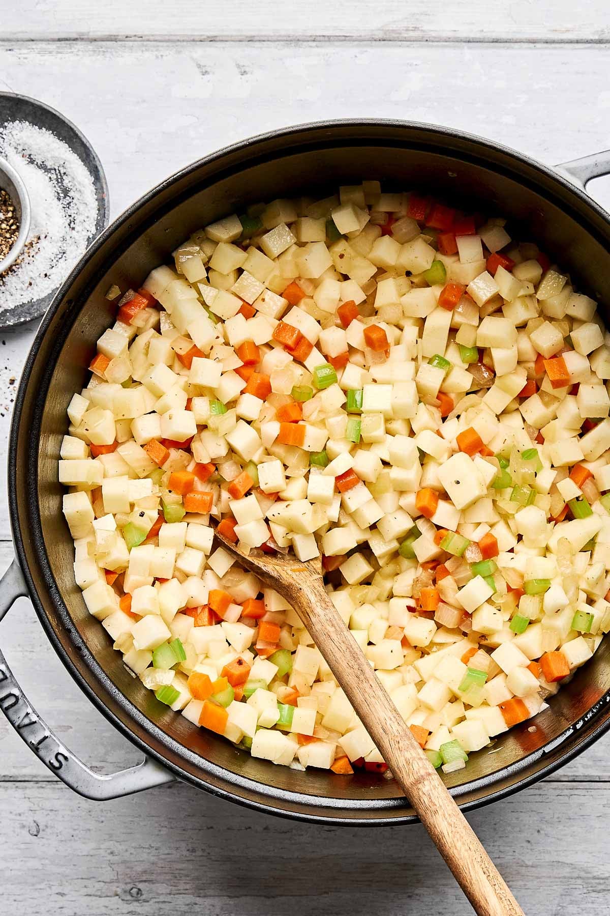 A large pot filled with diced vegetables, including potatoes, carrots, celery, and onions, being stirred with a wooden spoon; a small dish of salt and pepper sits nearby on a light surface.
