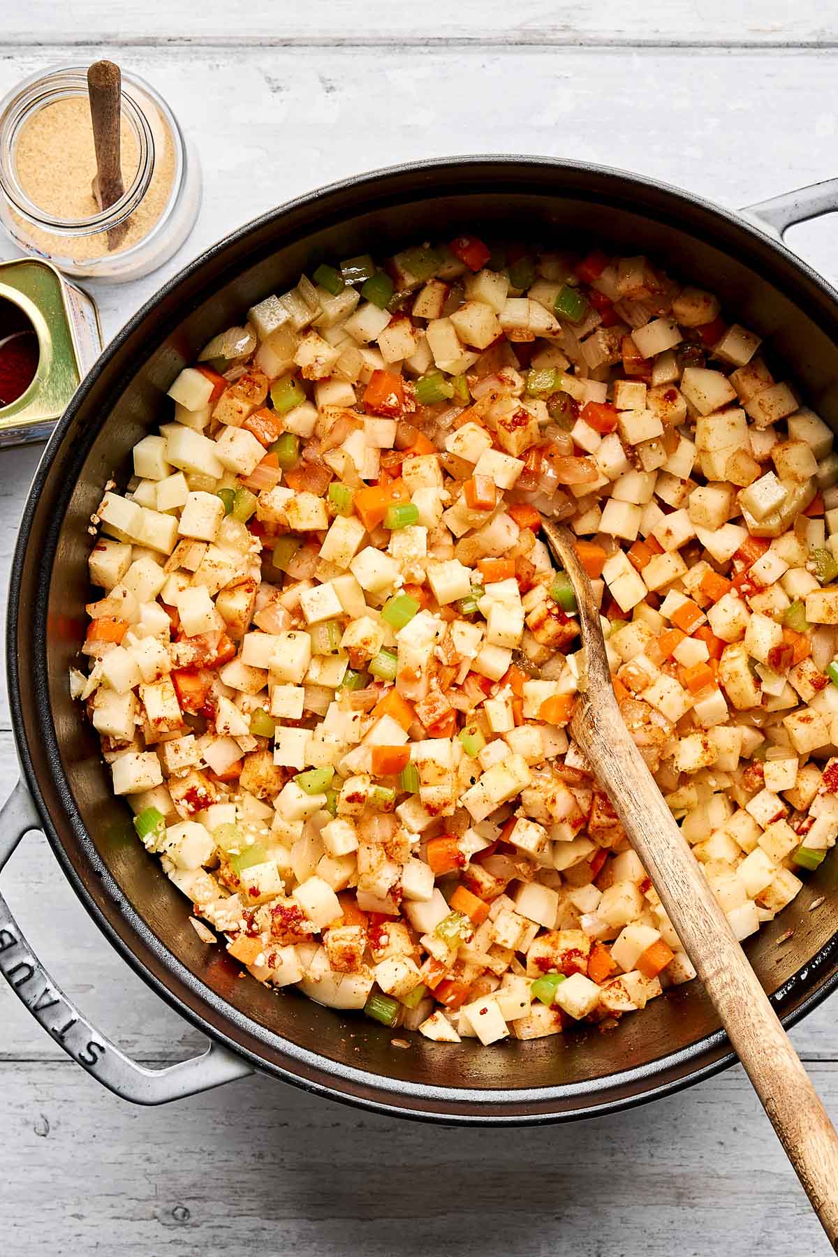 A pot filled with diced vegetables, including potatoes, carrots, and celery, being stirred with a wooden spoon. Jars of spices sit nearby on a light wooden surface.