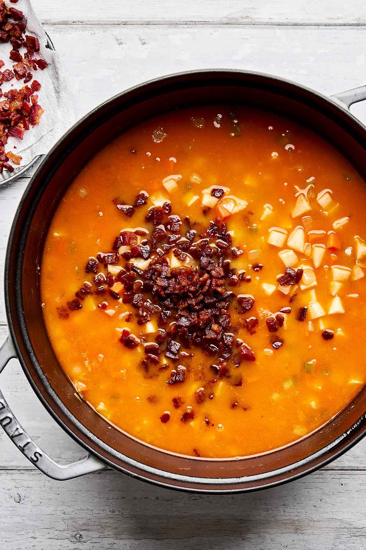 A large pot of orange-red broth with diced potatoes and a generous topping of crispy bacon bits, sitting on a light-colored wooden surface. Bacon pieces are also visible on a napkin nearby.