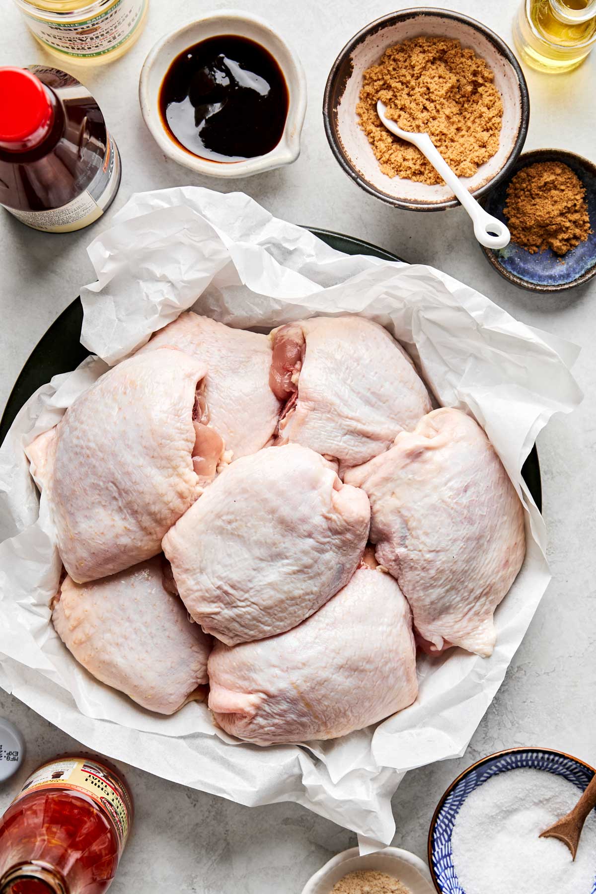 Raw chicken thighs are arranged in a bowl lined with white parchment paper, surrounded by small bowls containing brown sugar, soy sauce, and other liquid and powdered ingredients on a light countertop.