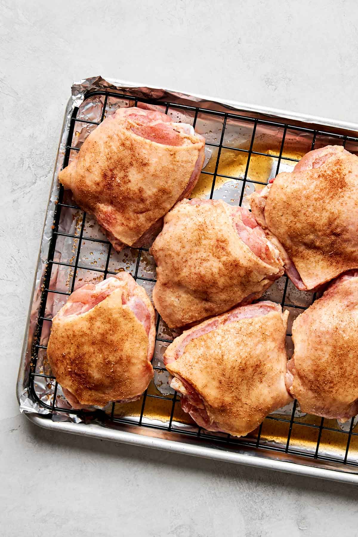 Six raw, seasoned chicken thighs are arranged on a wire rack placed over a foil-lined baking sheet. The chicken is sprinkled with a brown spice mix. The background is a light gray surface.