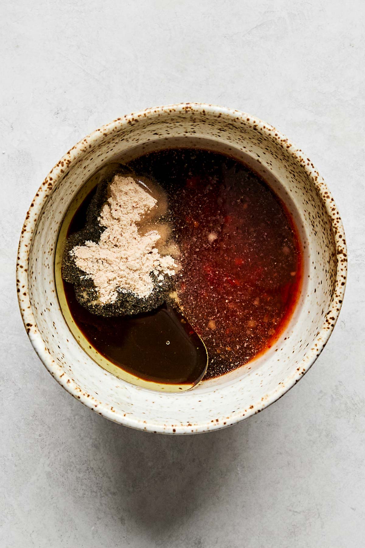A speckled ceramic bowl filled with a mixture of spices, dark liquid, and reddish sauce, sitting on a light gray surface.