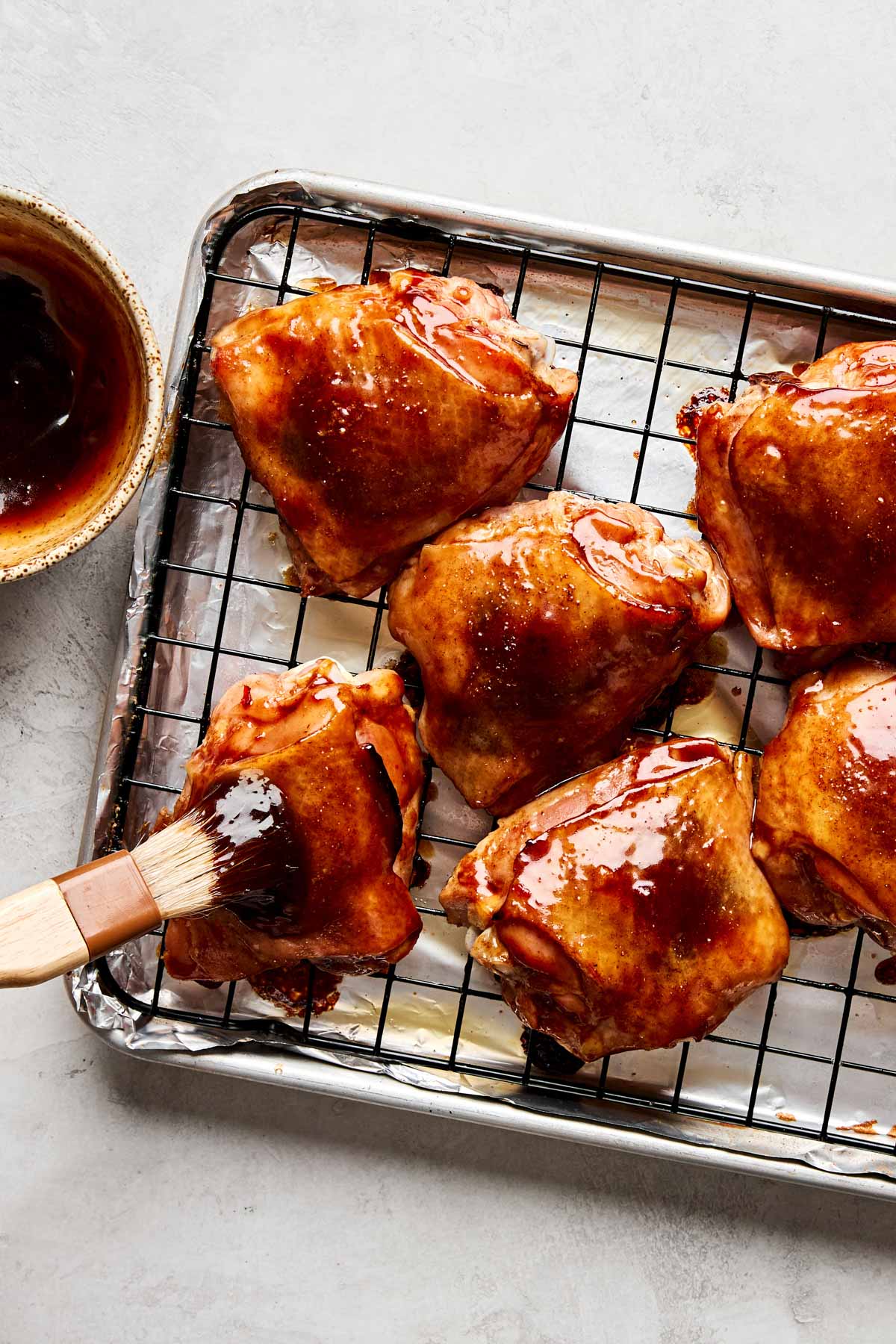 Six Peking chicken thighs on a wire rack over a baking sheet being brushed with a thick, glossy sauce; a bowl of sauce sits nearby.