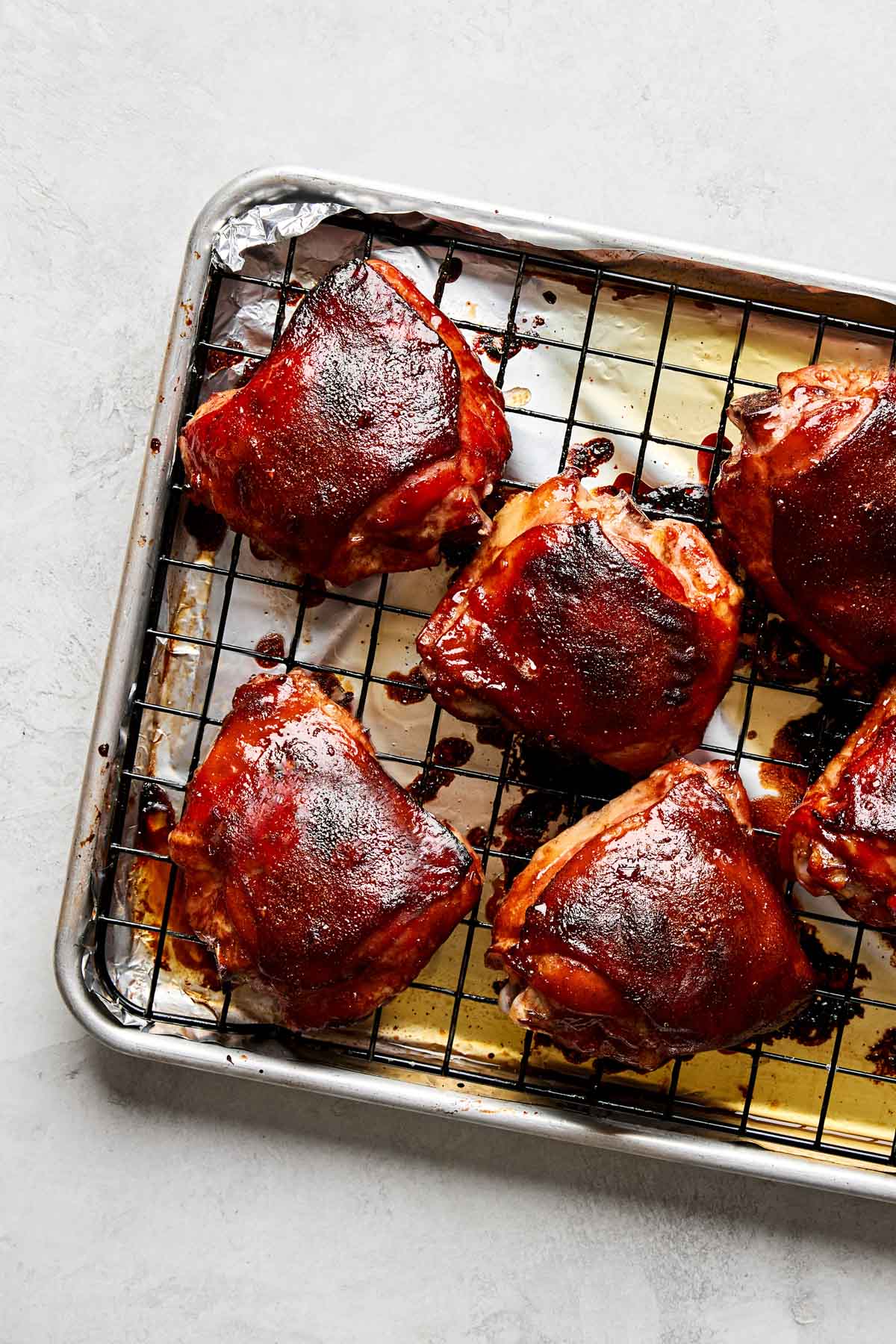 Six glazed, cooked Peking style chicken thighs rest on a wire rack over a baking sheet lined with foil. The chicken has a shiny, dark reddish-brown coating and appears juicy and well-roasted.