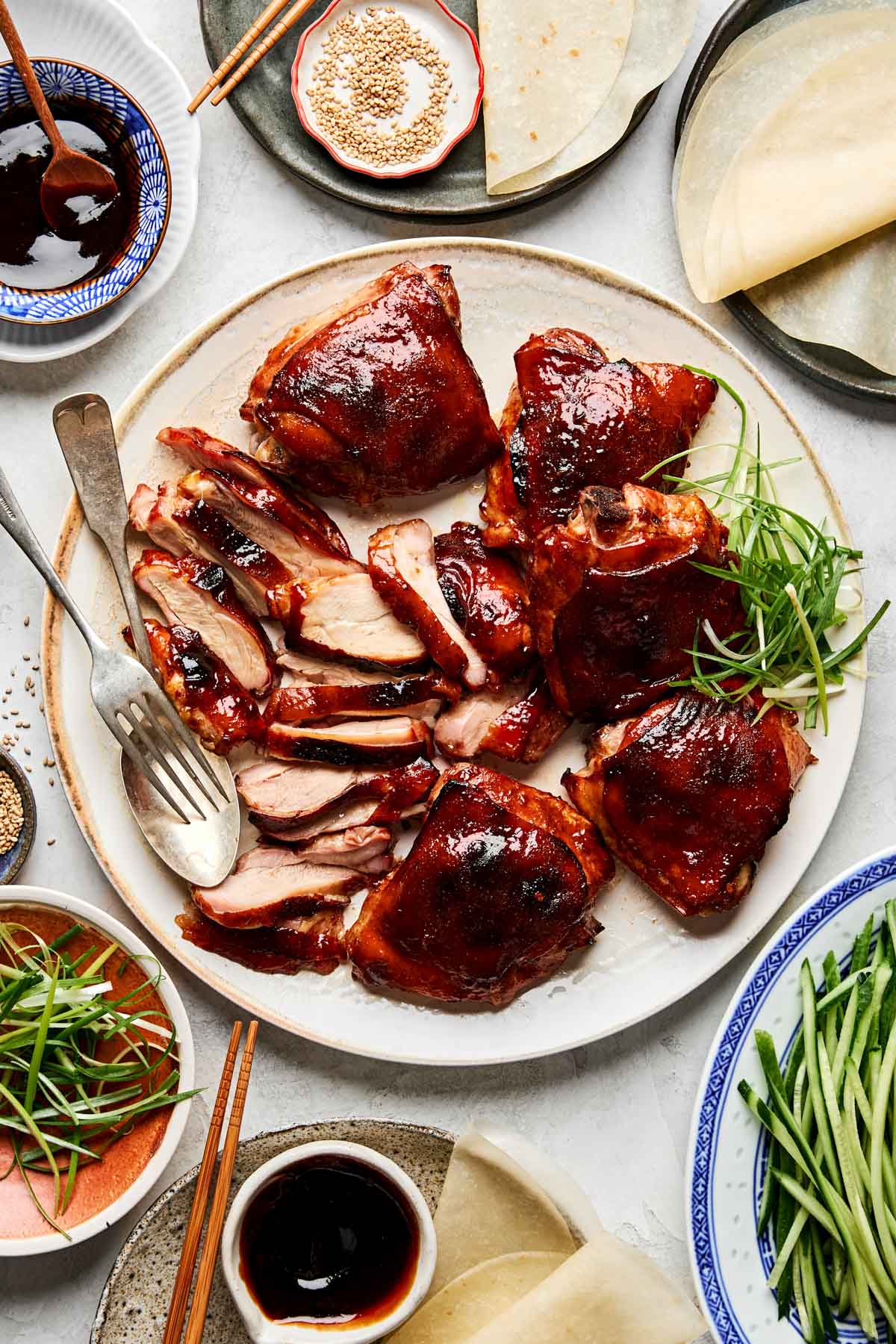 A plate of sliced Chinese roast Peking chicken thighs with crispy skin, surrounded by thin pancakes, sliced green onions, cucumber, hoisin sauce, and sesame seeds, ready for assembling Peking duck wraps.