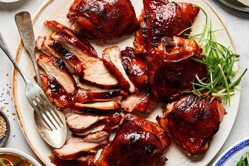 A plate with glazed, roasted Peking chicken thighs, some whole and some sliced, garnished with fresh green onions. A serving fork and spoon are placed beside the chicken on the plate.