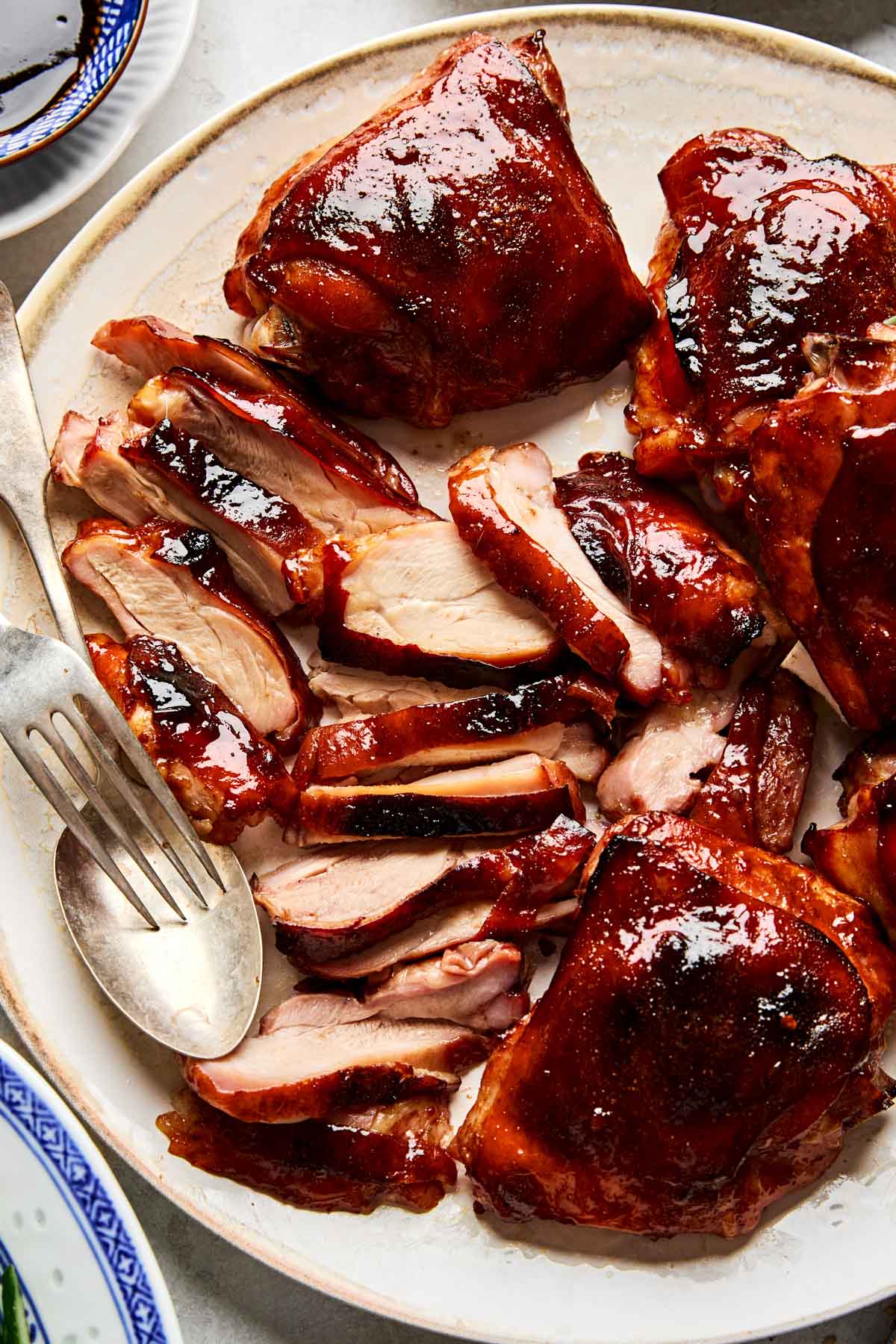 A plate of glazed, roasted Peking style chicken thighs, some whole and some sliced, with a shiny, caramelized exterior. A serving fork rests on the plate beside the chicken.