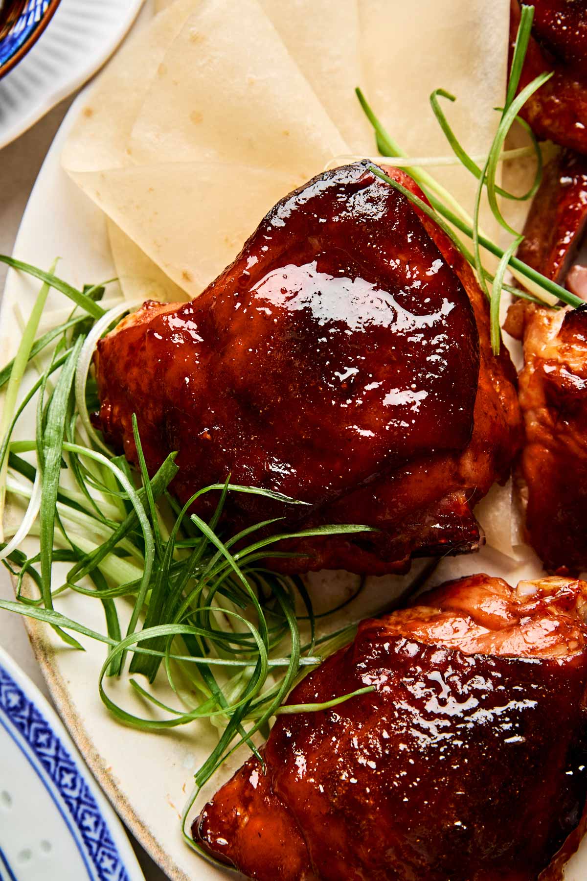 Glossy, dark glazed pieces of cooked Peking chicken are served on a plate with thinly sliced green onions and several pale, round pancakes or wrappers in the background.