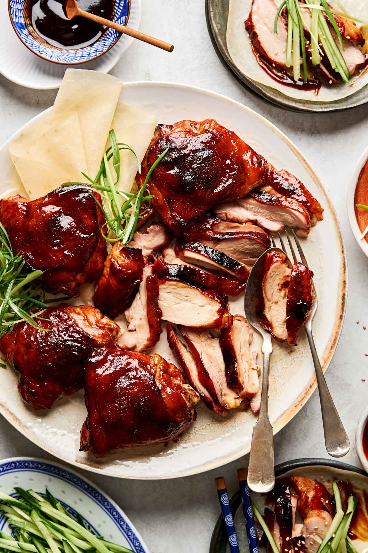 A plate of sliced roast Peking chicken with glossy, crispy skin, served with green onion strips and thin pancakes. Small plates and dishes with sauces and more duck slices surround the main platter.