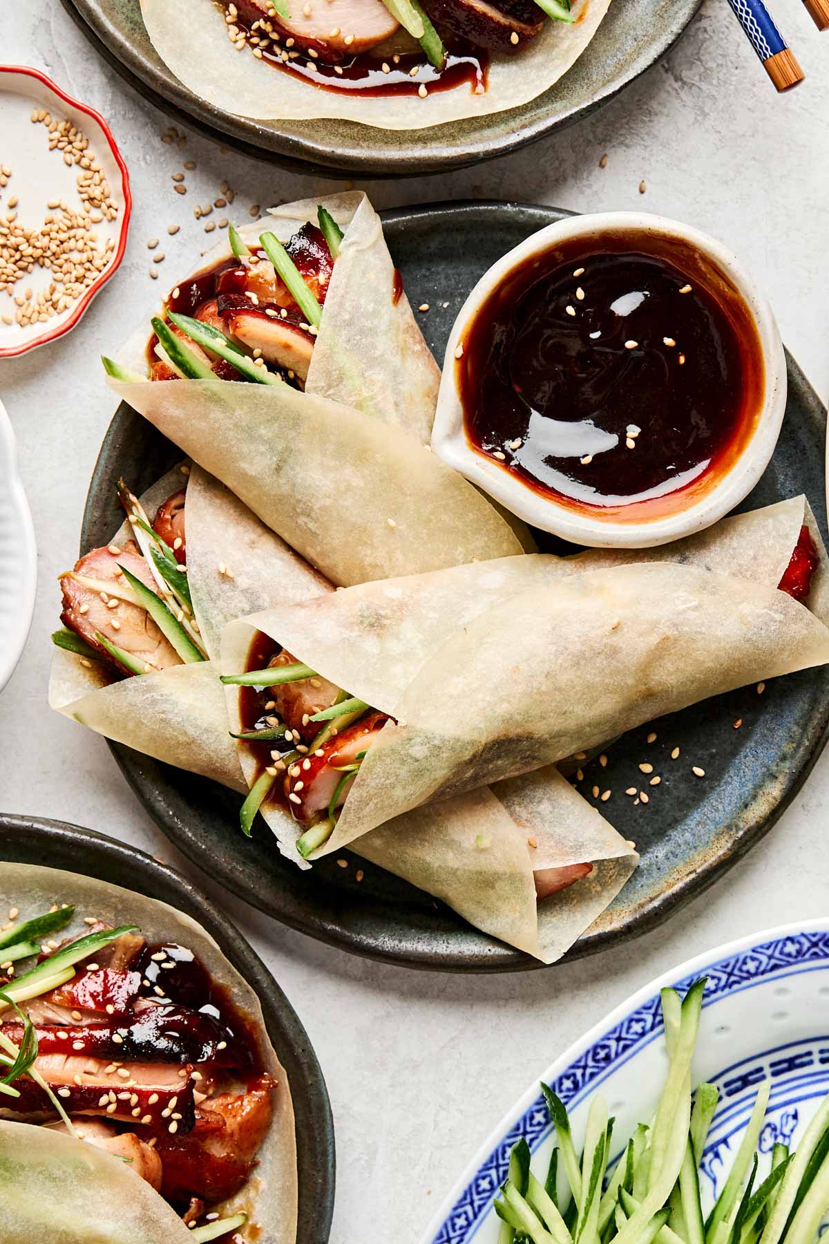 Three Chinese-style pancakes filled with sliced Peking chicken, cucumber, and hoisin sauce are arranged on a plate with a small bowl of dipping sauce. Some sesame seeds are sprinkled on top, and cucumber strips are nearby.
