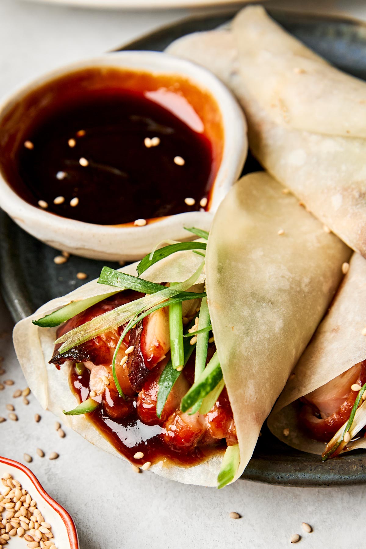 Two Chinese-style pancakes filled with sliced Peking chicken thighs, cucumber, and green onions, served on a dark plate with a bowl of hoisin sauce topped with sesame seeds.